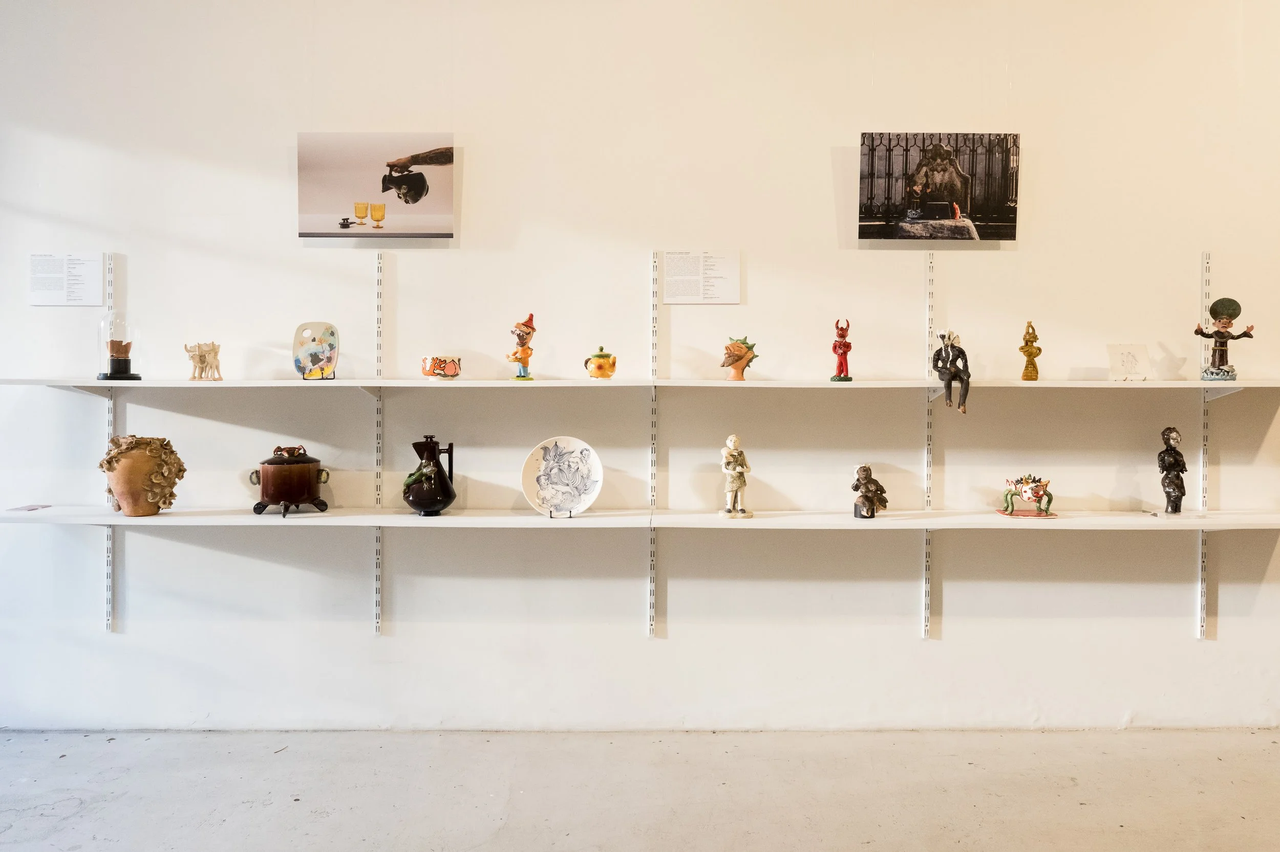 White gallery wall with three shelves displaying various small art sculptures and two framed photographs above the shelves.