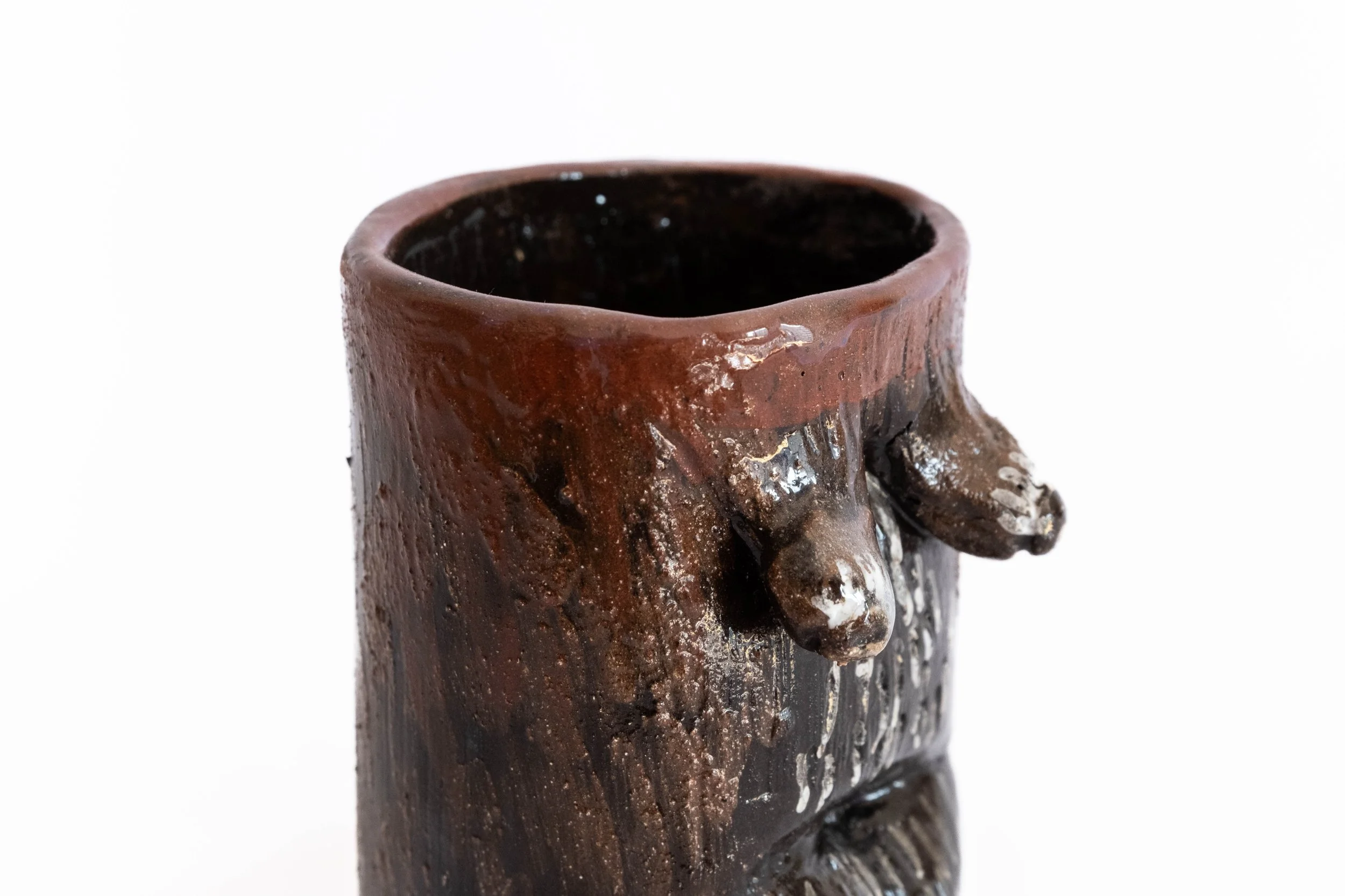 Close-up of a brown ceramic mug with a textured, carved design and a handle shaped like an animal's paws, against a plain white background.