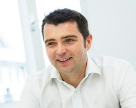 A smiling man with dark hair wearing a white shirt in a bright office setting.
