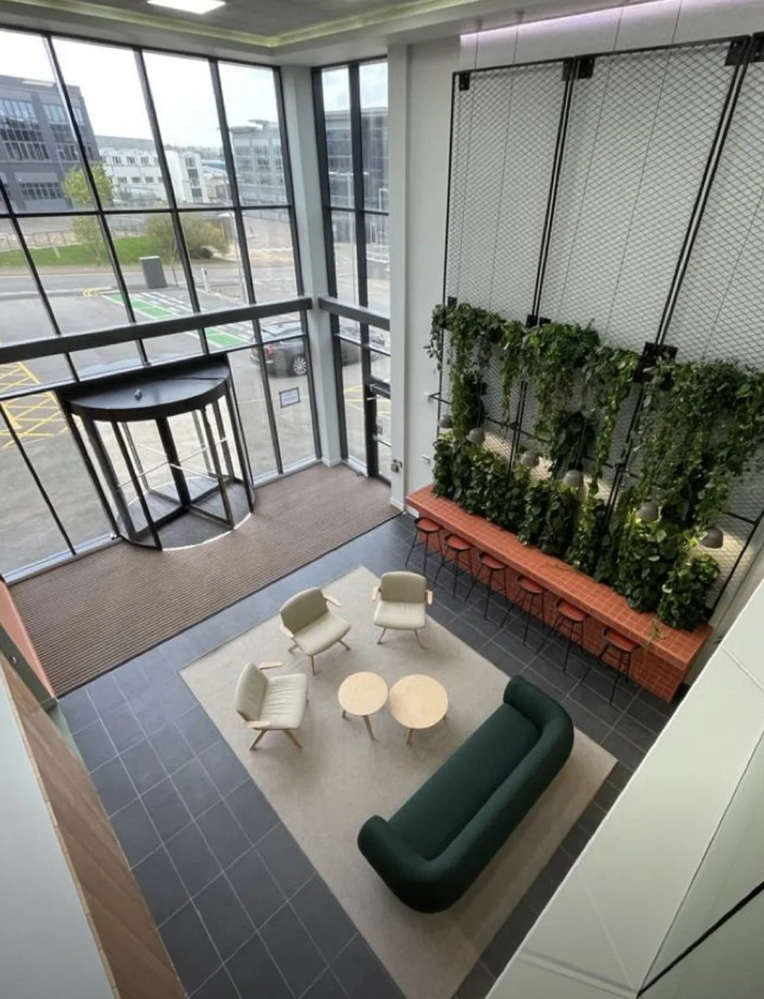 Indoor lounge area with large floor-to-ceiling windows, beige chairs, a green sofa, and a wall with green plants and hanging lights.