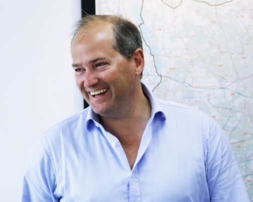 A man smiling in front of a map on a wall, wearing a light blue collared shirt.