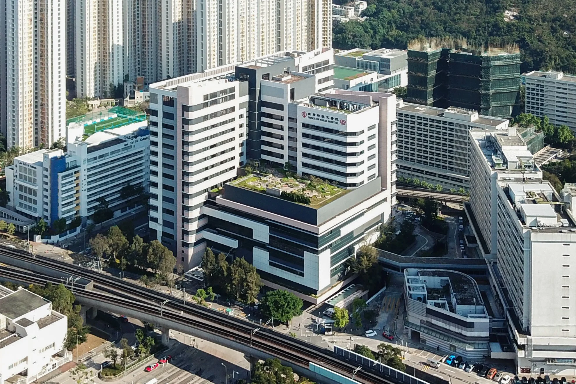 A large medical building with multiple floors, rooftop greenery, and a hospital sign, surrounded by high-rise buildings and city infrastructure.