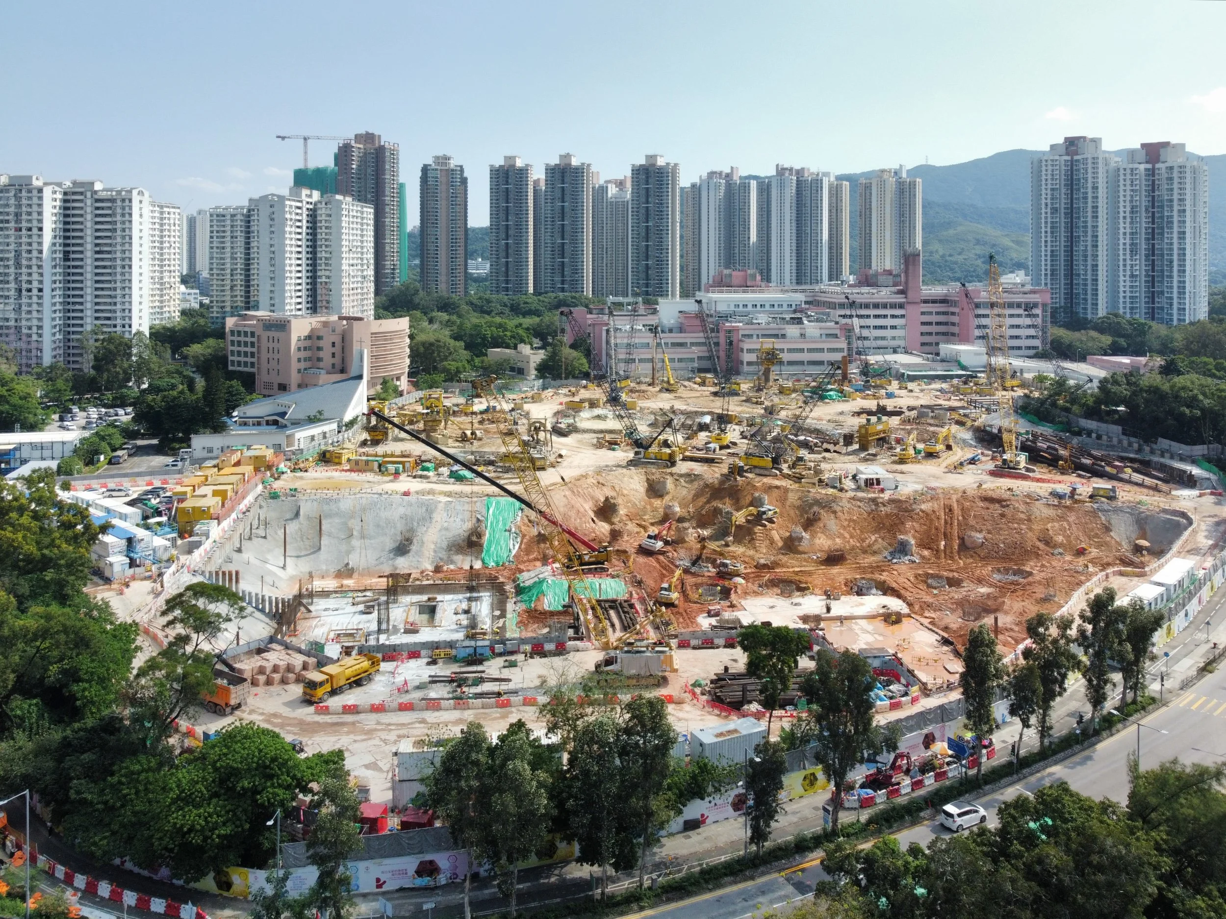 A large construction site with multiple cranes and machinery, surrounded by high-rise buildings in an urban area of Hong Kong.