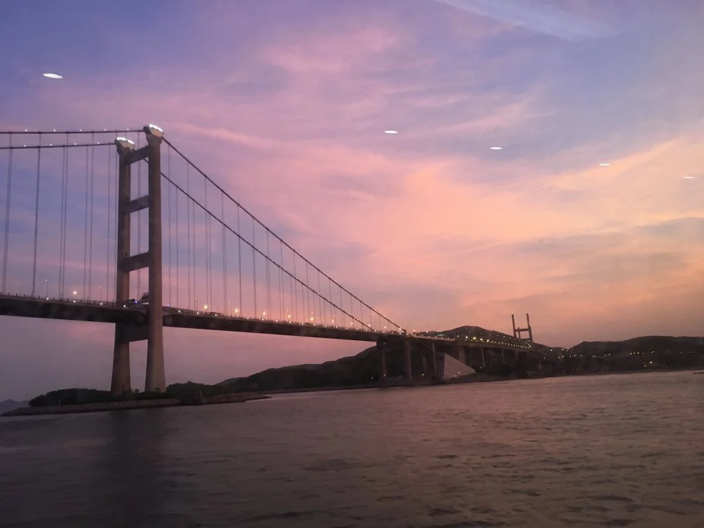 The Tsing Ma suspension bridge at sunset with pink and purple sky, spanning over water with hills in the background.