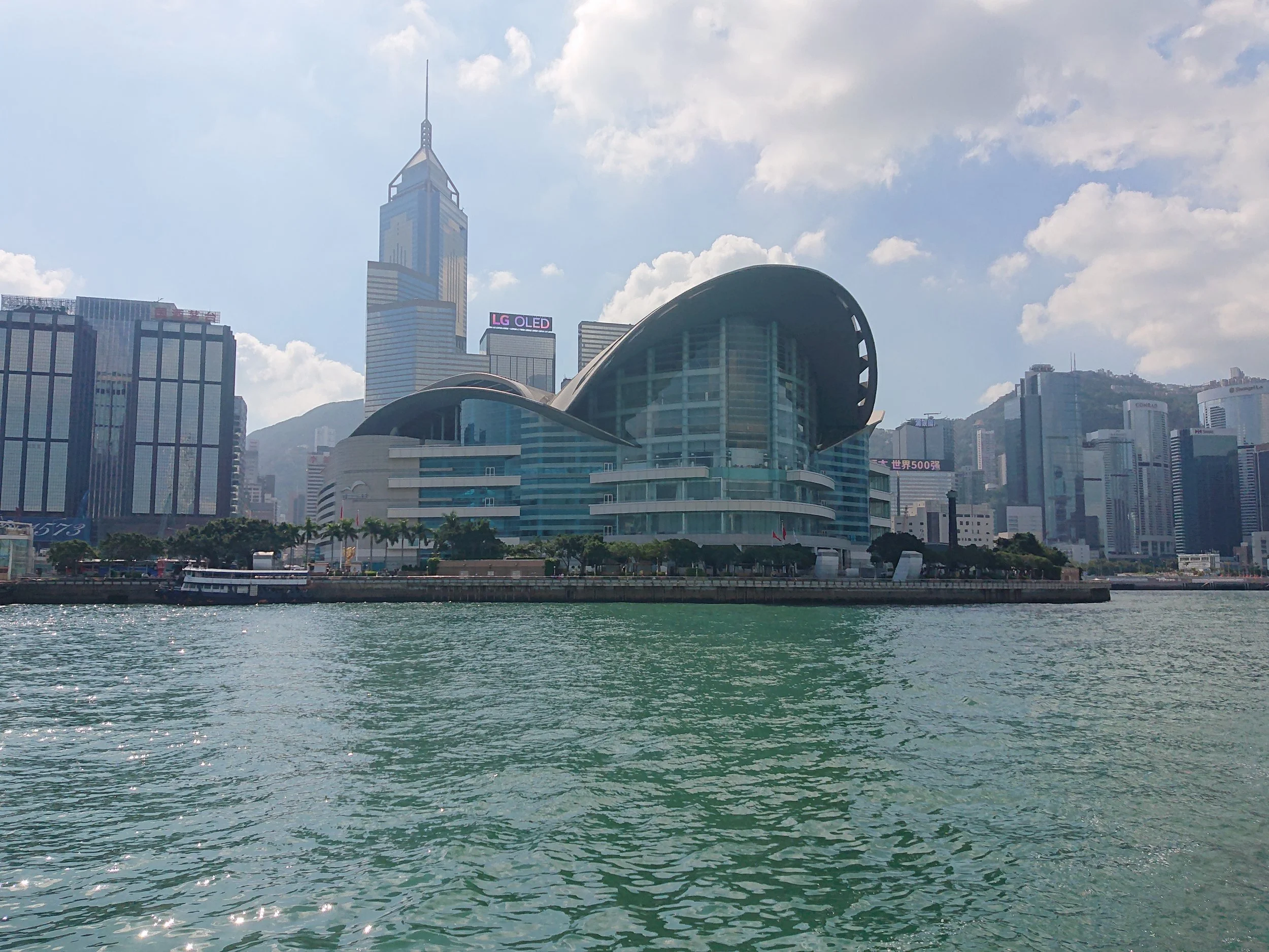 Hong Kong island skyline with view of the Hong Kong Convention and Exhibition Centre on the Victoria Harbour  a. unique curved glass building, tall skyscrapers in background, and Victoria Peak behind.