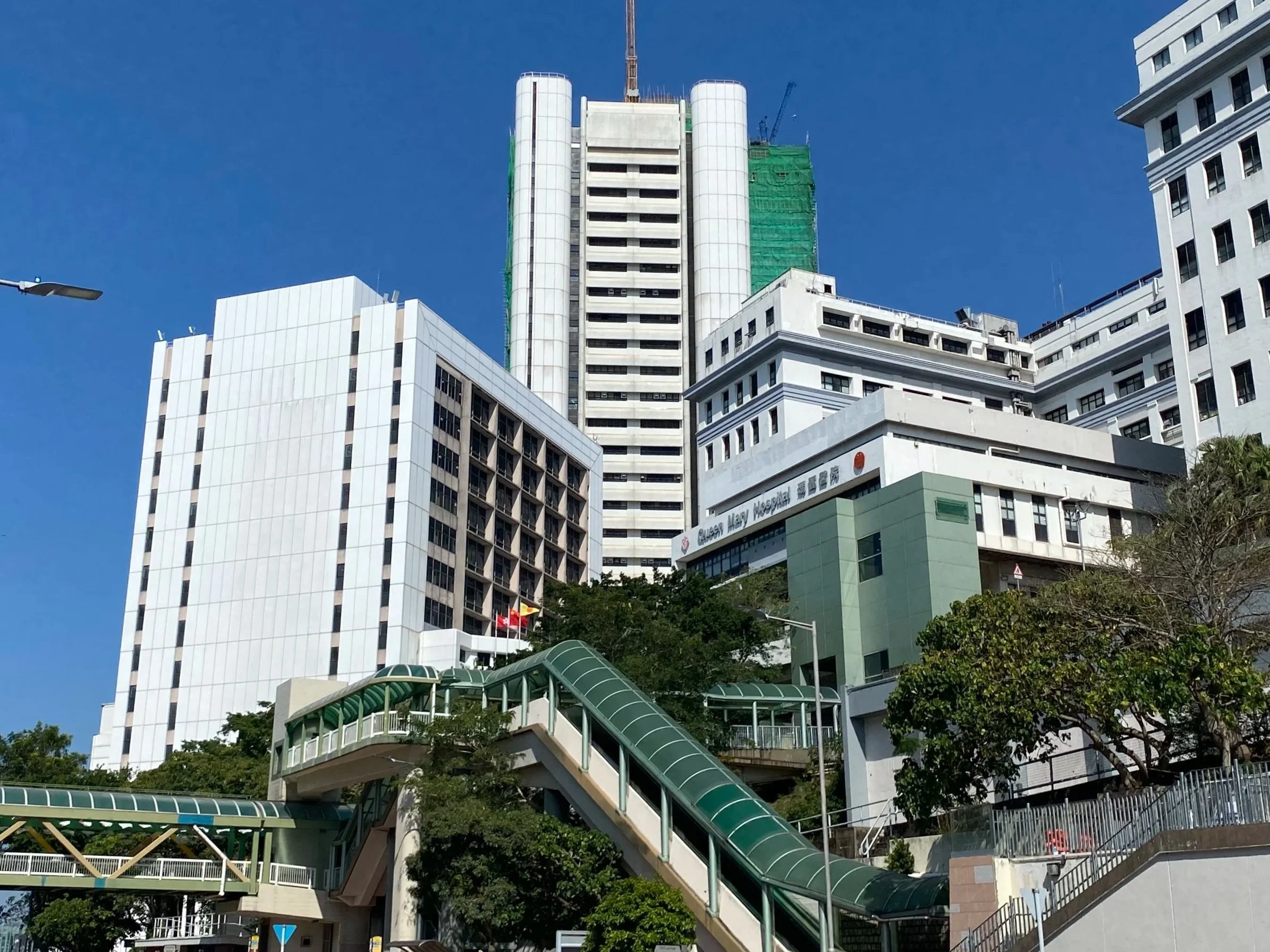 Tall white and green hospital buildings with a skybridge and trees in front.