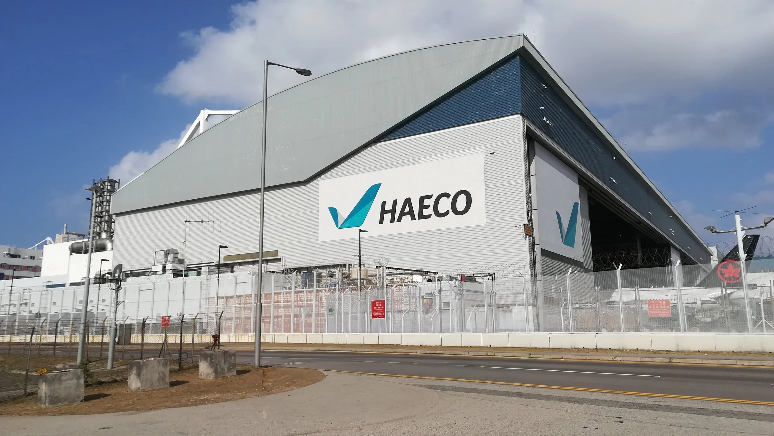 Large industrial building with the HAЕCO logo on the side, surrounded by a chain-link fence and barbed wire, under a partly cloudy sky.