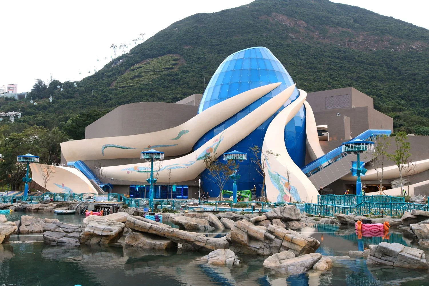 Ocean Park's futuristic building with a large blue glass dome and white structural elements, set against a green mountain background, with a body of water and rocks in the foreground.
