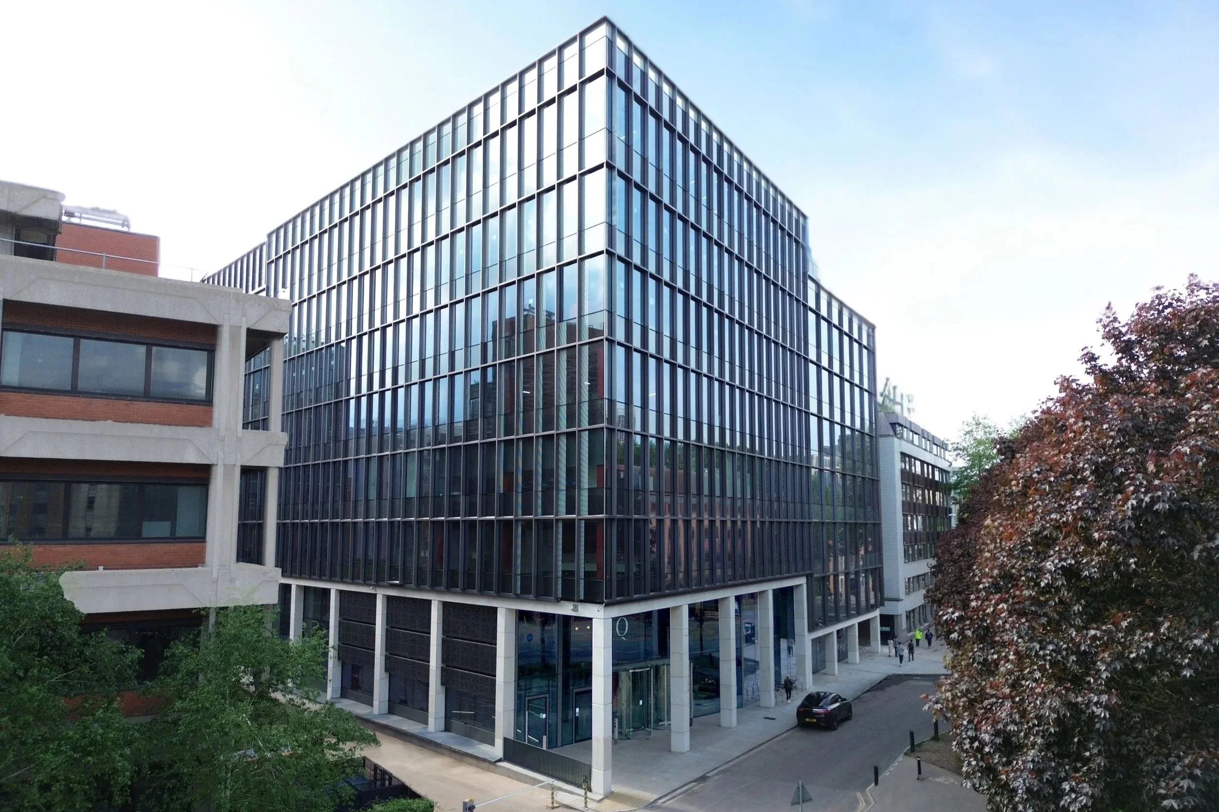 Modern multi-story office building with glass facade reflecting the sky, adjacent to a smaller building with brick and concrete exterior, and trees on the street below.