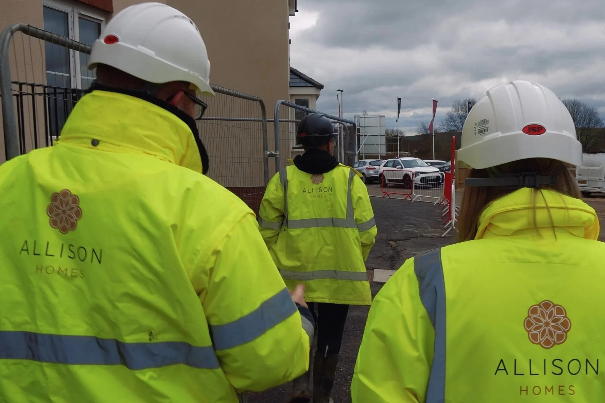 Construction workers wearing yellow high-visibility jackets and white safety helmets at a worksite with parked cars and cloudy sky in the background.