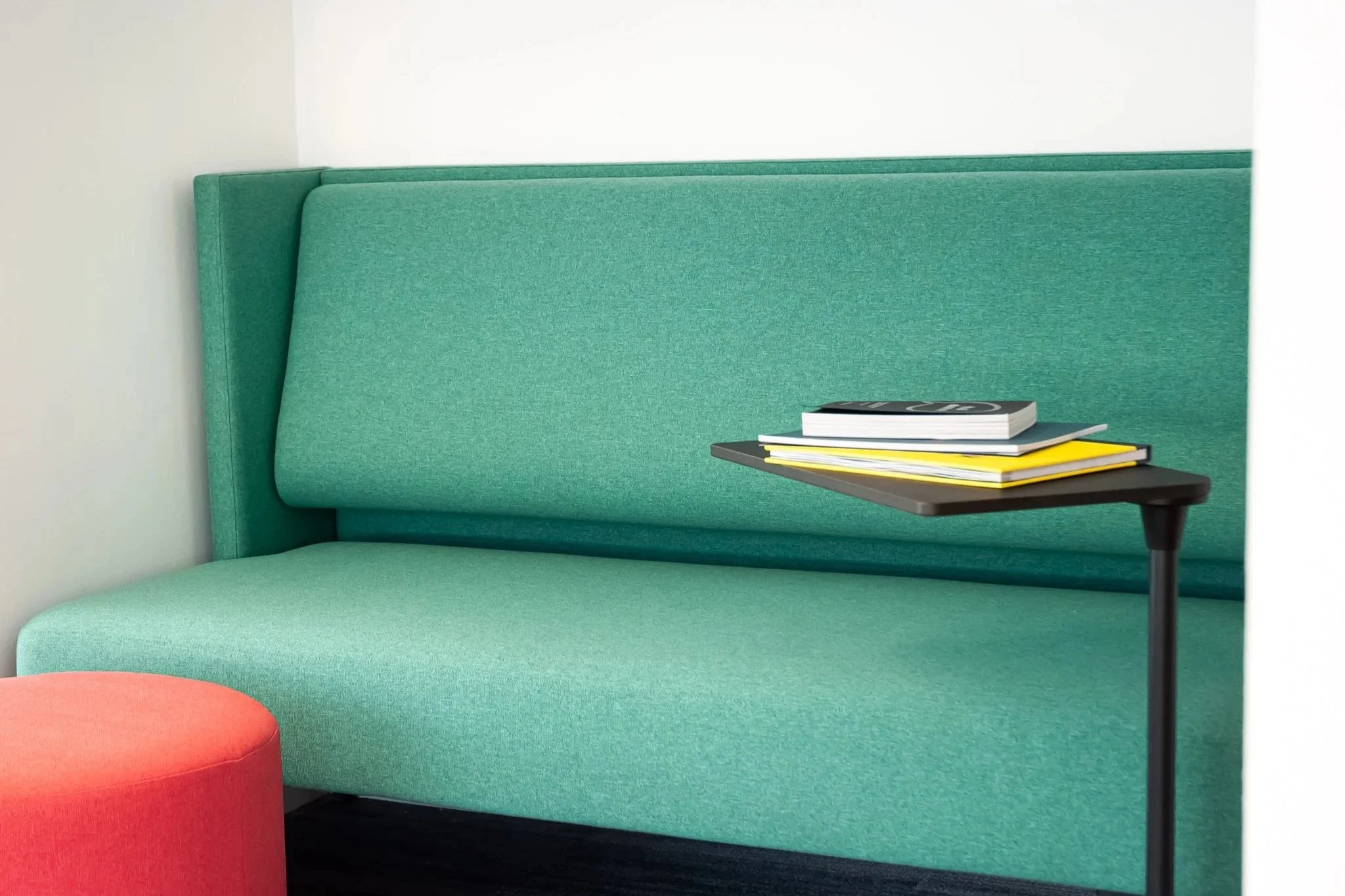 A minimalist seating area with a green upholstered booth and a small black side table holding a few magazines. A red round ottoman is partially visible in the foreground.