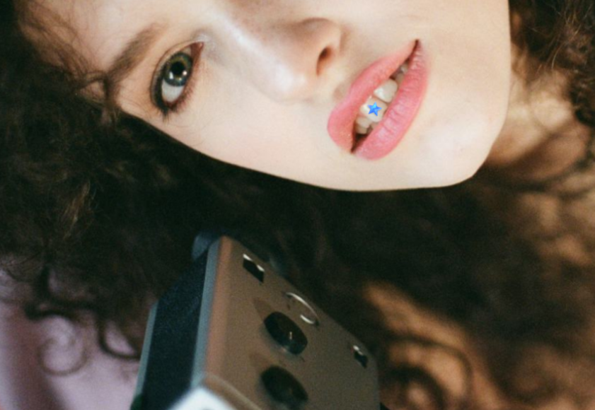 Close-up of a young woman with curly hair, partially lying down, holding a camera, with her head tilted and tongue sticking out, revealing a star crystal on her tooth.