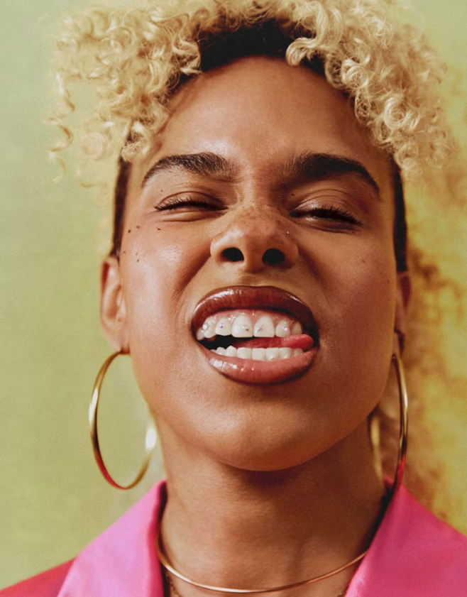 A woman with curly blonde hair, wearing large gold hoop earrings and a pink jacket, making a playful face with her tongue sticking out and eyes squinting.