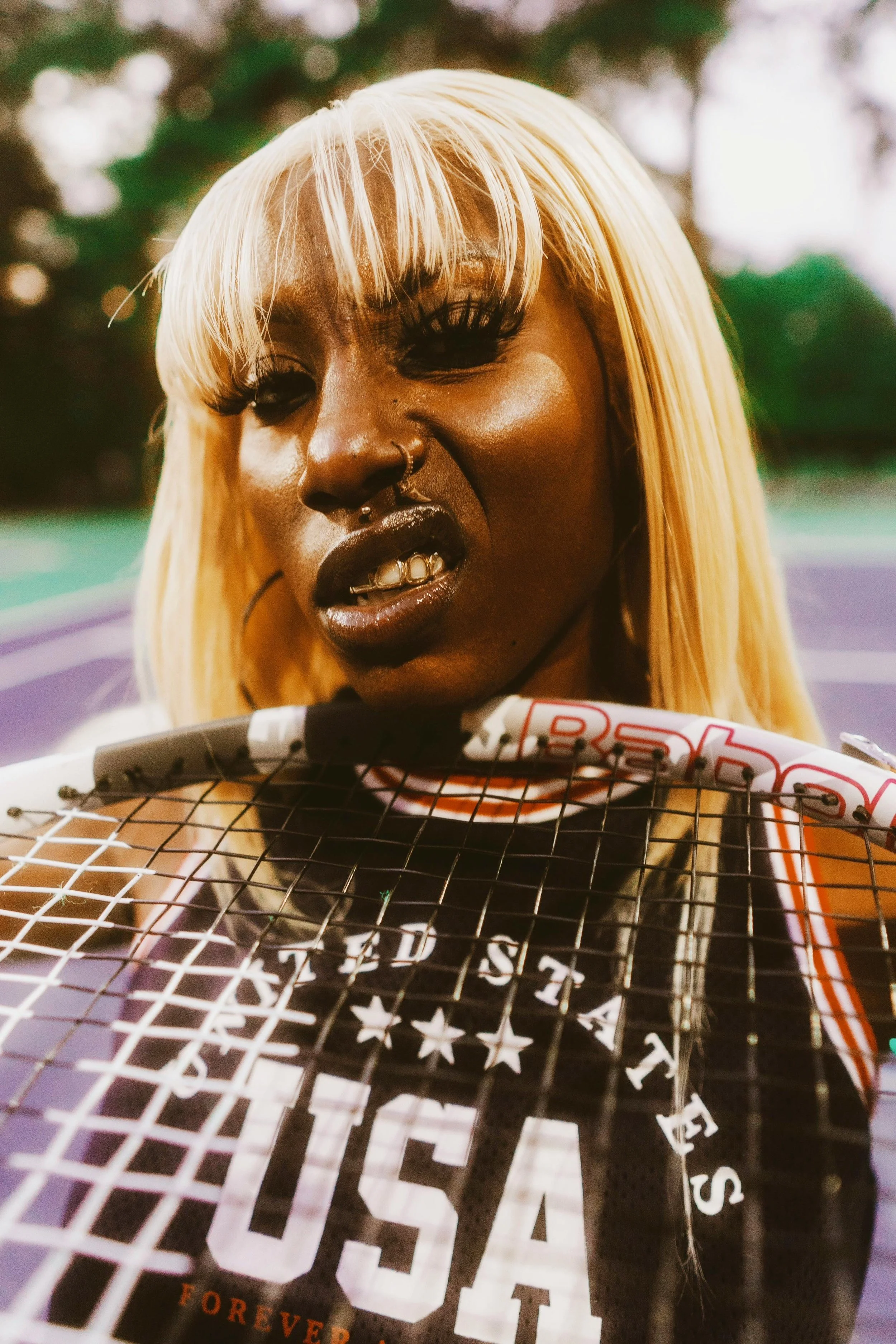 A woman with blonde hair, a set of gold grillz, and elaborate makeup, gripping a tennis racket near her face. She wears a sports jersey and has multiple facial piercings. The background is blurred with what looks like an outdoor sports court.
