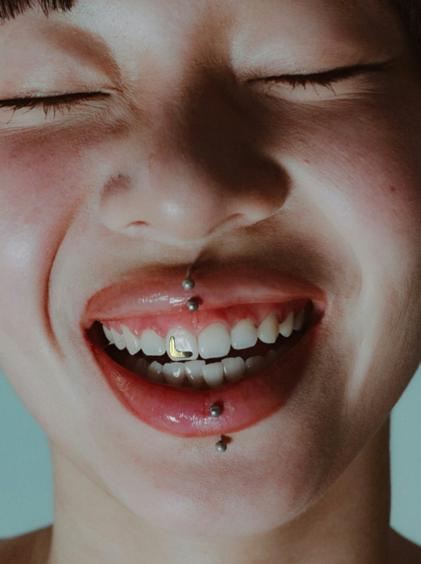 Close-up of a smiling person with a gold charm on the front tooth, multiple facial piercings, including a septum piercing, a lip piercing, and a piercing on the tongue.