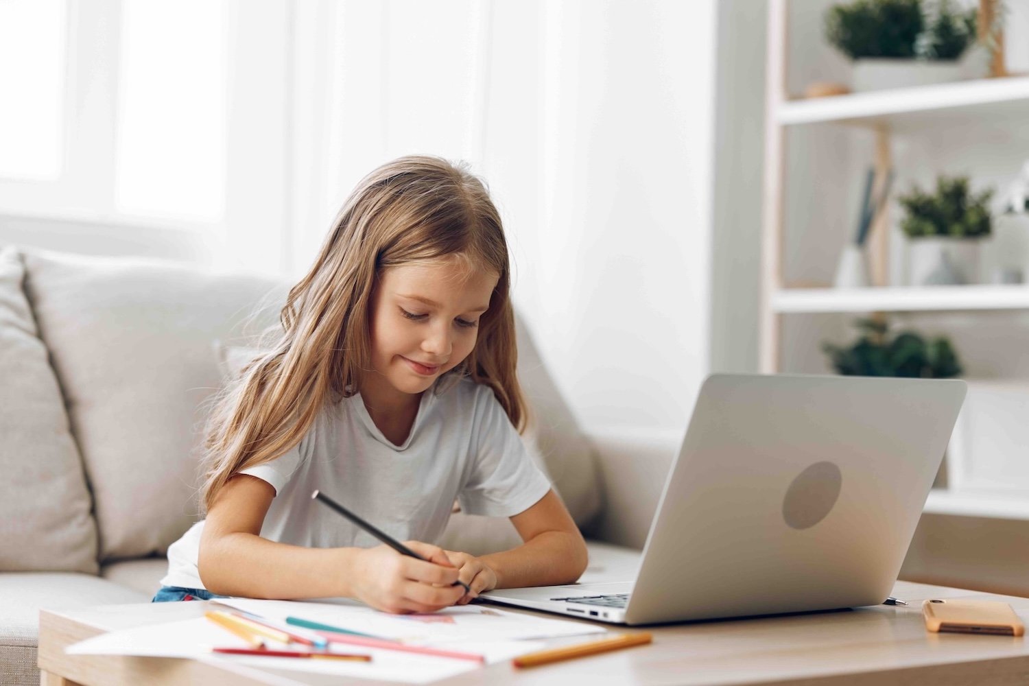 Primary school girl doing an online tutoring lesson at home, writing with a pencil beside a laptop