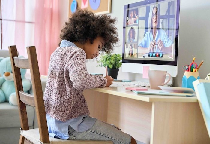 Primary school child taking an online group tutoring lesson, writing at a desk while the teacher talks on screen