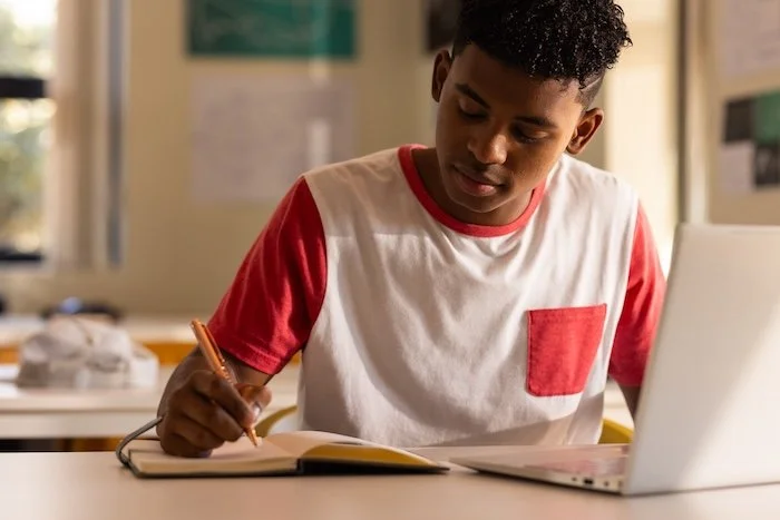 Secondary school student writing notes beside a laptop during an online tutoring lesson.