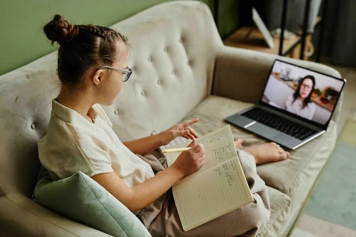 Primary school girl taking an online tutoring lesson from home, writing notes in a notebook while listening to her teacher on a laptop