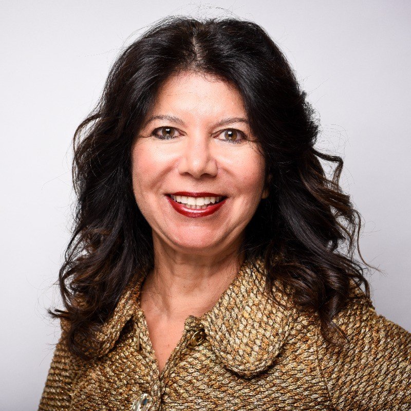 Close-up of a woman with curly hair, wearing glasses, hoop earrings, and a striped shirt, smiling indoors.
