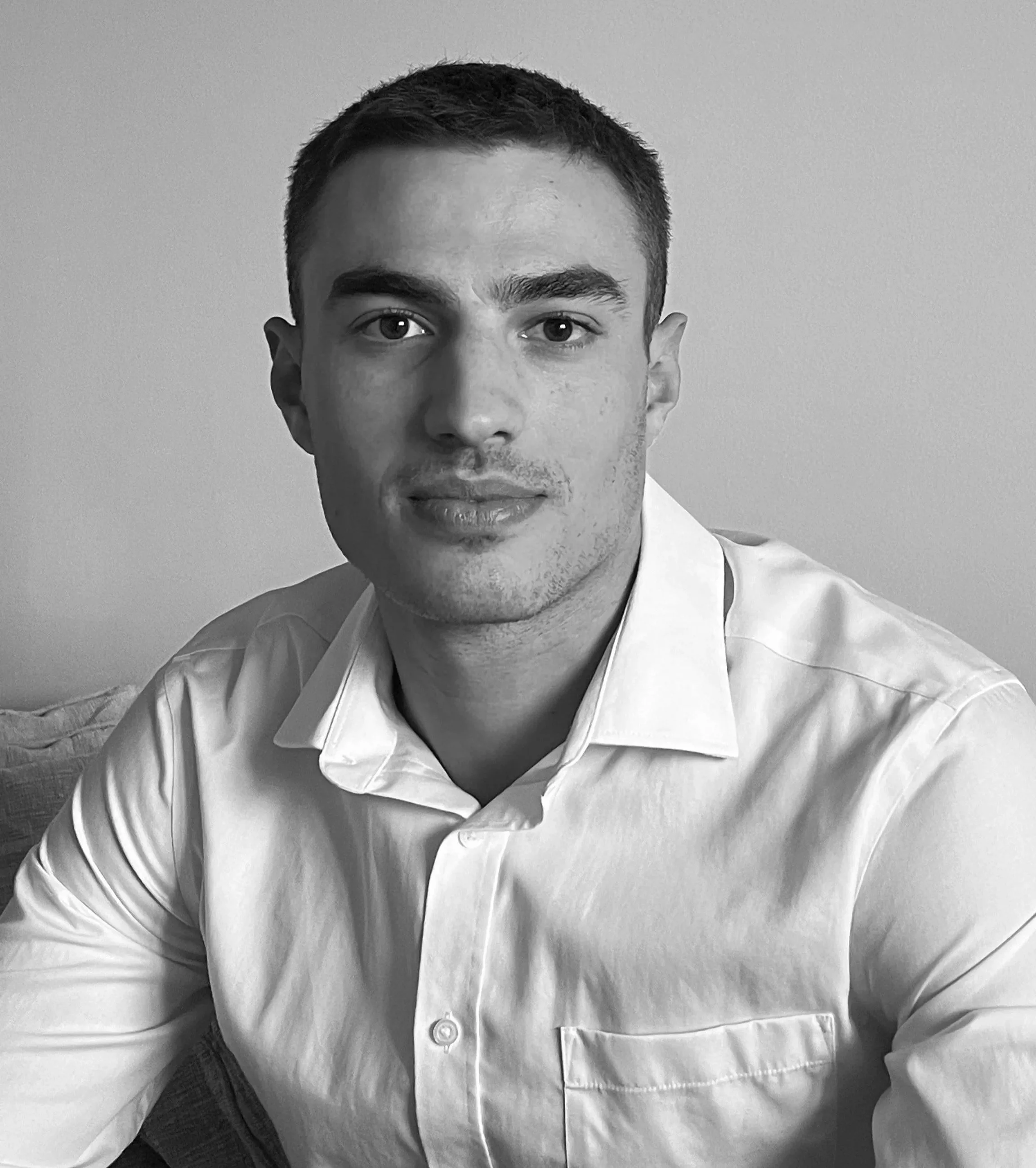 Black and white portrait of a young man with short hair, wearing a white button-up shirt, sitting indoors with a neutral background.