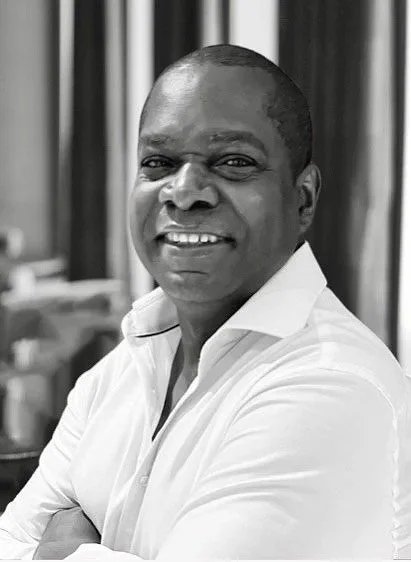 A smiling man with short hair, wearing a white shirt, standing in an indoor setting with blurred background.