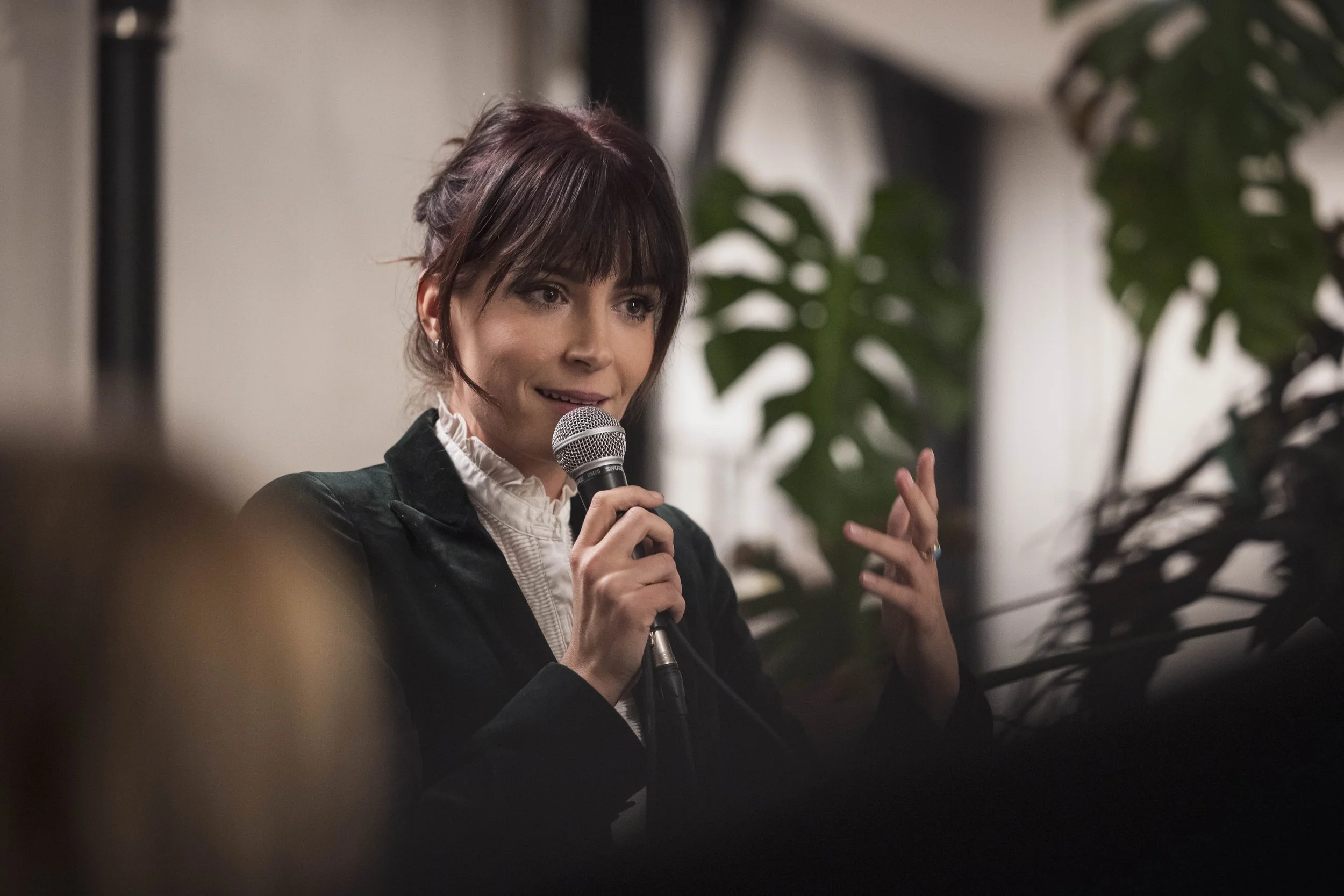 Une femme parle dans un microphone lors d'une discussion ou d'une conférence, dans un environnement intérieur avec des plantes en arrière-plan.