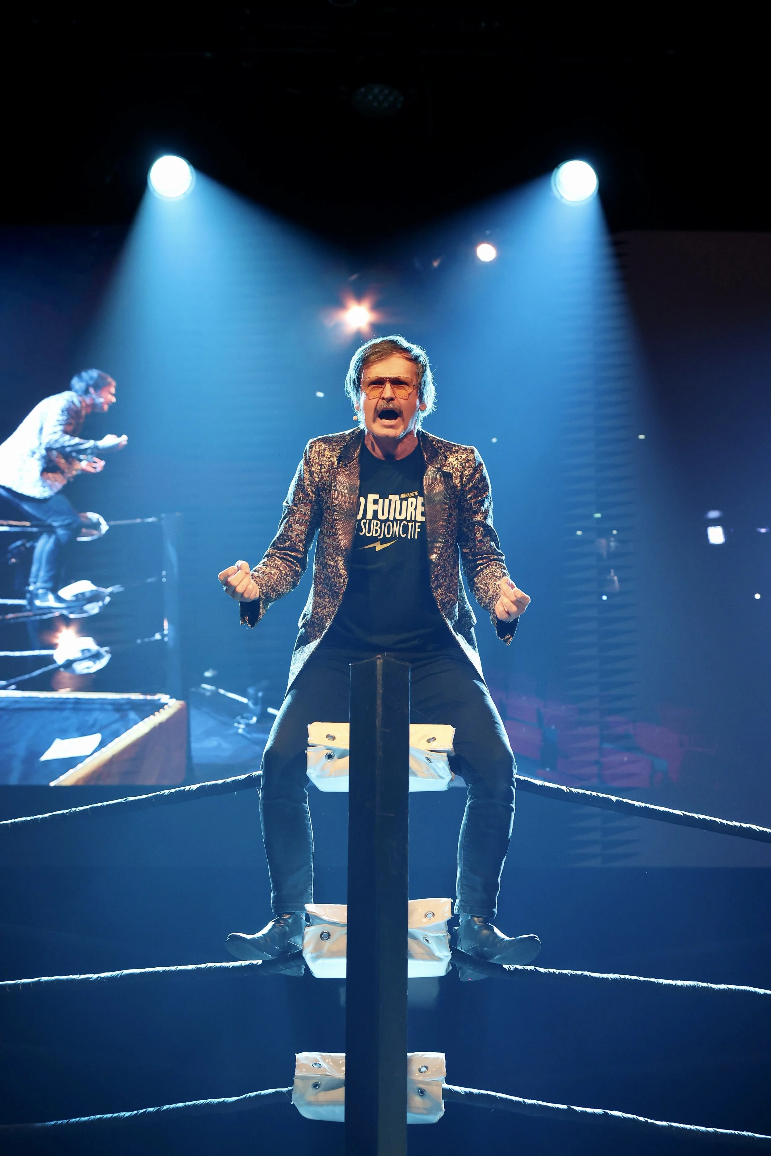 Un homme debout au centre d'un ring de lutte, criant, avec des lumières impressionnantes et un autre en arrière-plan, dans une scène de performance ou de spectacle.