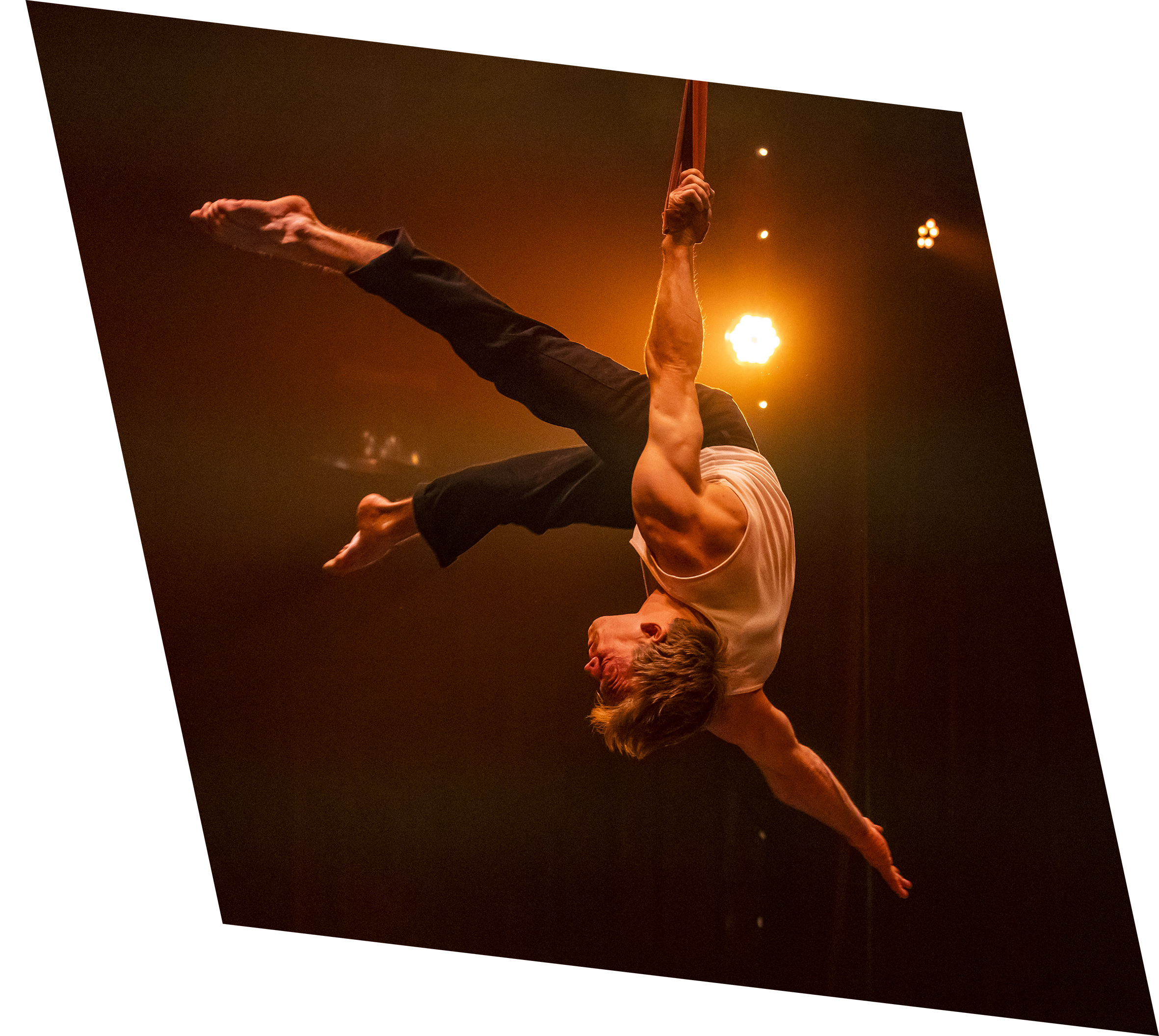 Un homme en équilibre suspendu dans les airs, tenant une corde ou une barre, lors d'une performance acrobatique ou artistique, sous un éclairage chaud.