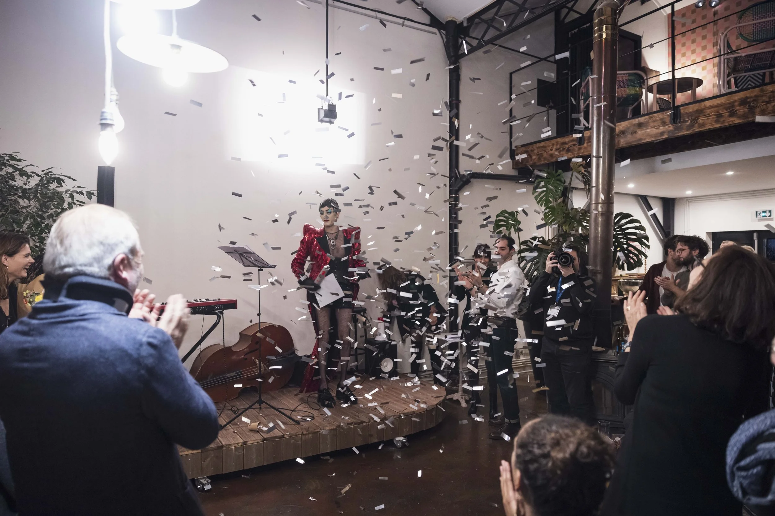 Une scène de célébration avec un musicien sur scène, des confettis tombant, des personnes applaudissant et prenant des photos lors d'un événement intérieur.