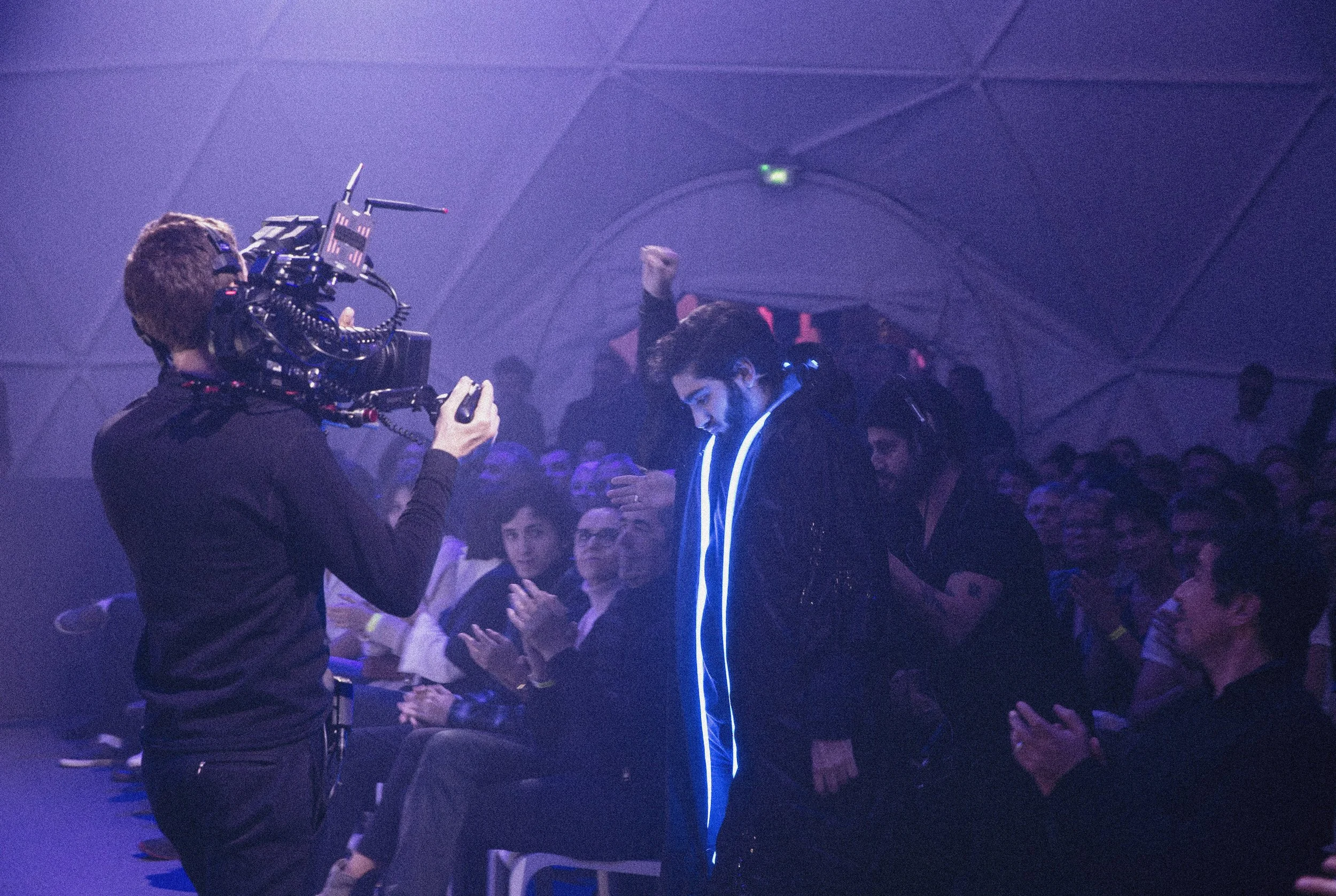Un homme portant une veste noire avec des lignes lumineuses bleues est en train de faire une présentation ou une performance dans une scène sombre, entouré d'un public qui l'applaudit. Un cameraman filme la scène.