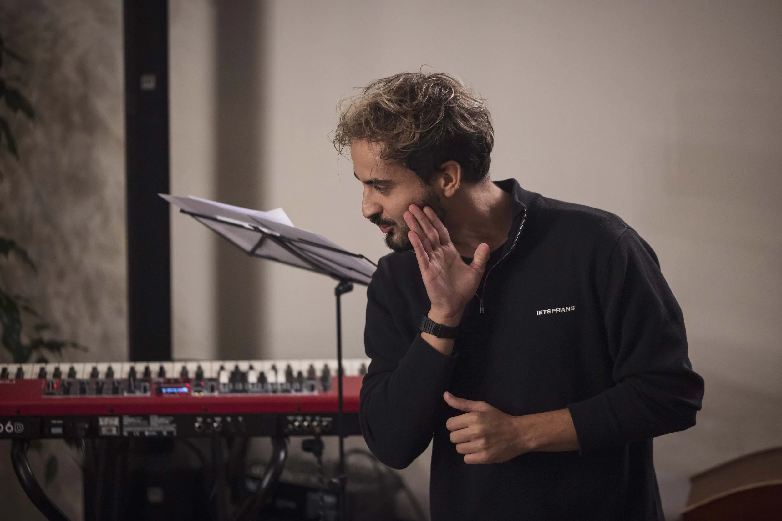 Un homme avec des cheveux bouclés et une barbe, portant un sweat noir, est en train de chanter ou de parler en public dans une pièce, près d'un clavier électronique.