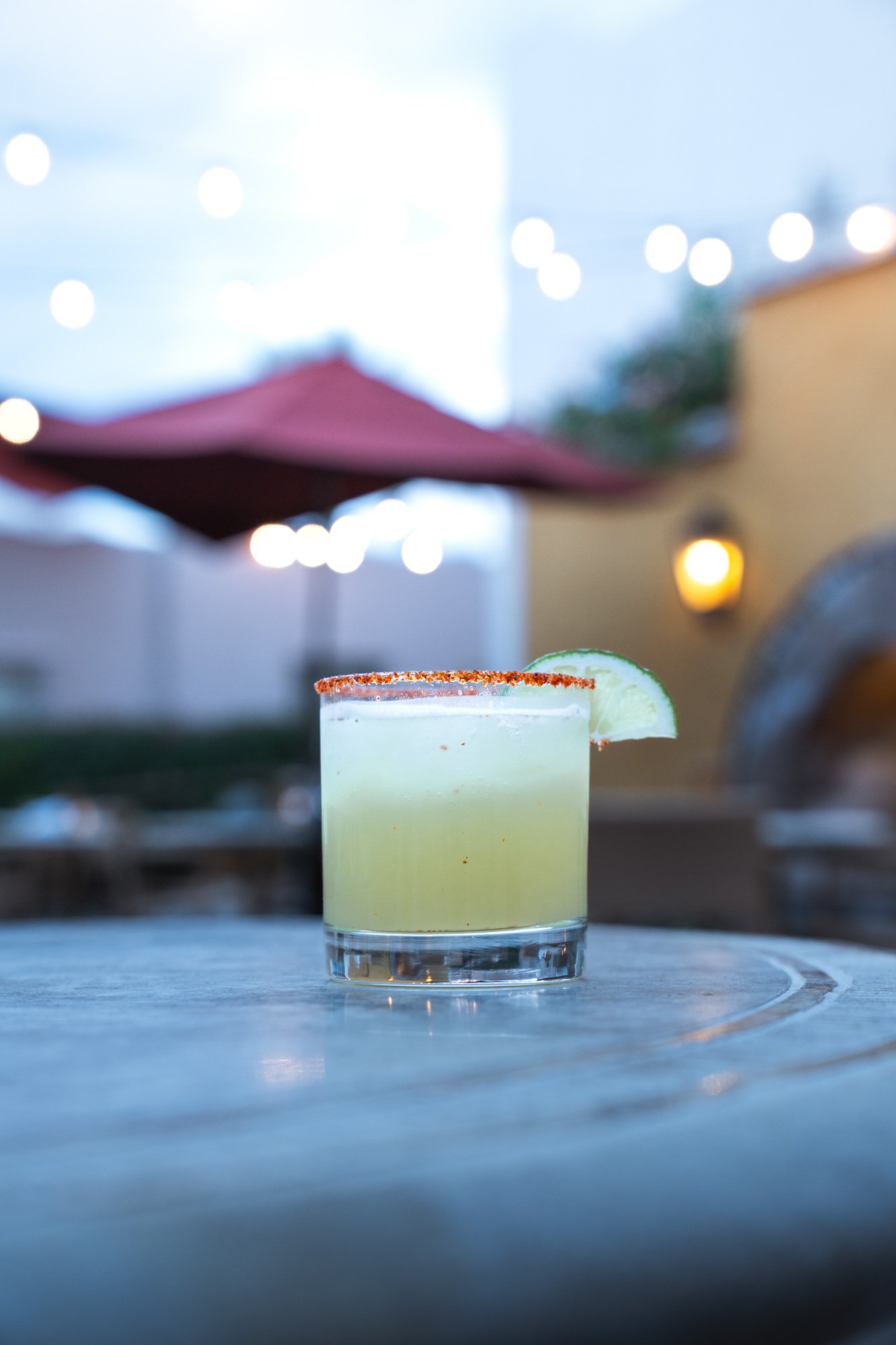 A lime margarita cocktail in a glass with a salted rim and a lime wedge garnish, placed on a wooden table outdoors in the evening.