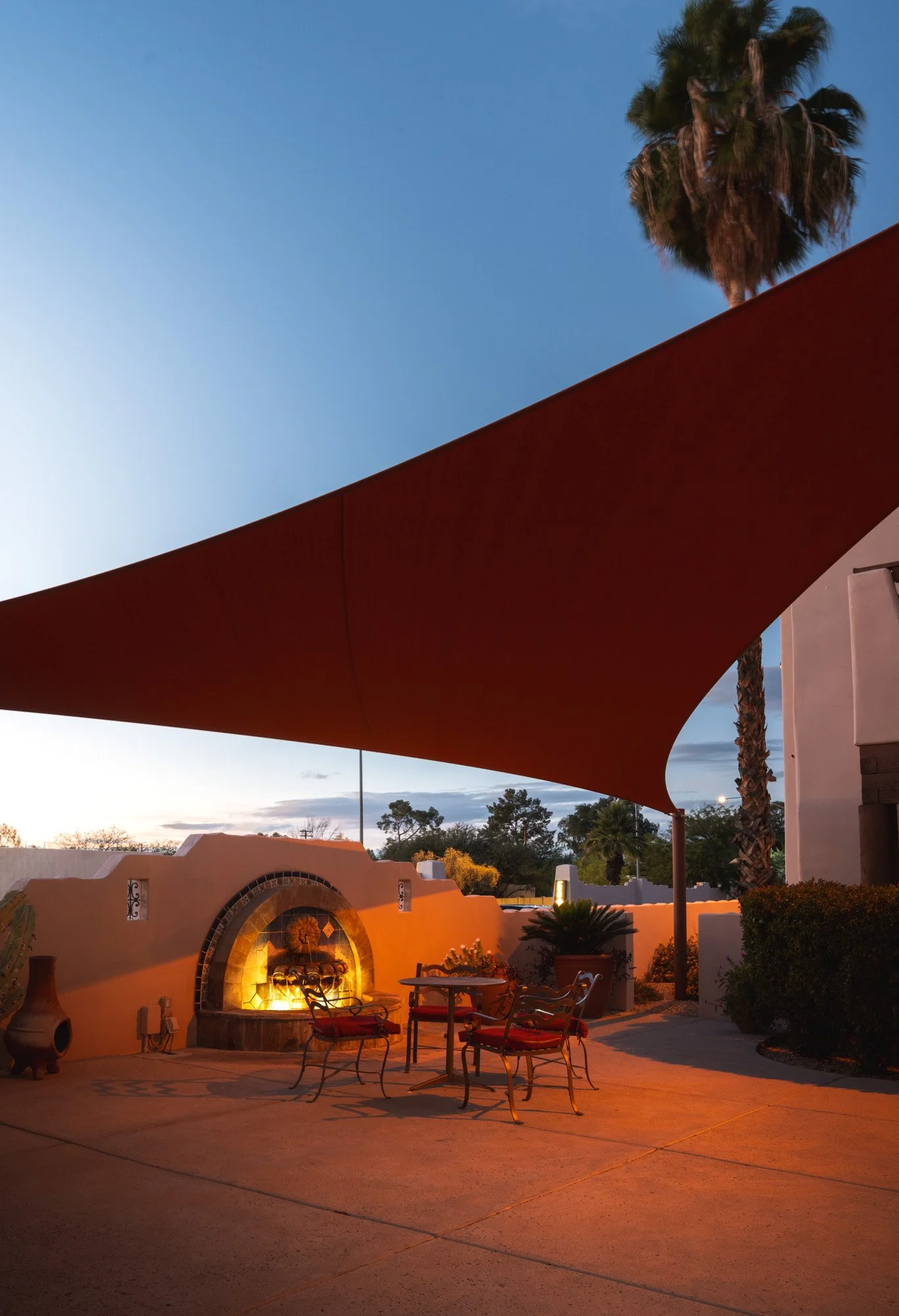 An outdoor patio with a large orange shade sail, a fire feature with warm lighting, a round glass table, and four metal chairs with red cushions. There are potted plants, a palm tree, and a beige wall with decorative tile accents, set against a backdrop of trees and a sunset sky.