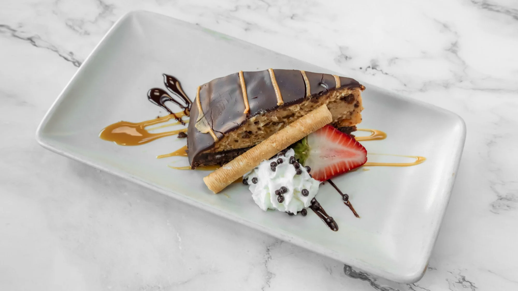 Slice of layered dessert with chocolate glaze, caramel drizzle, whipped cream, sliced strawberry, cookie stick, and chocolate pearls on a white plate.