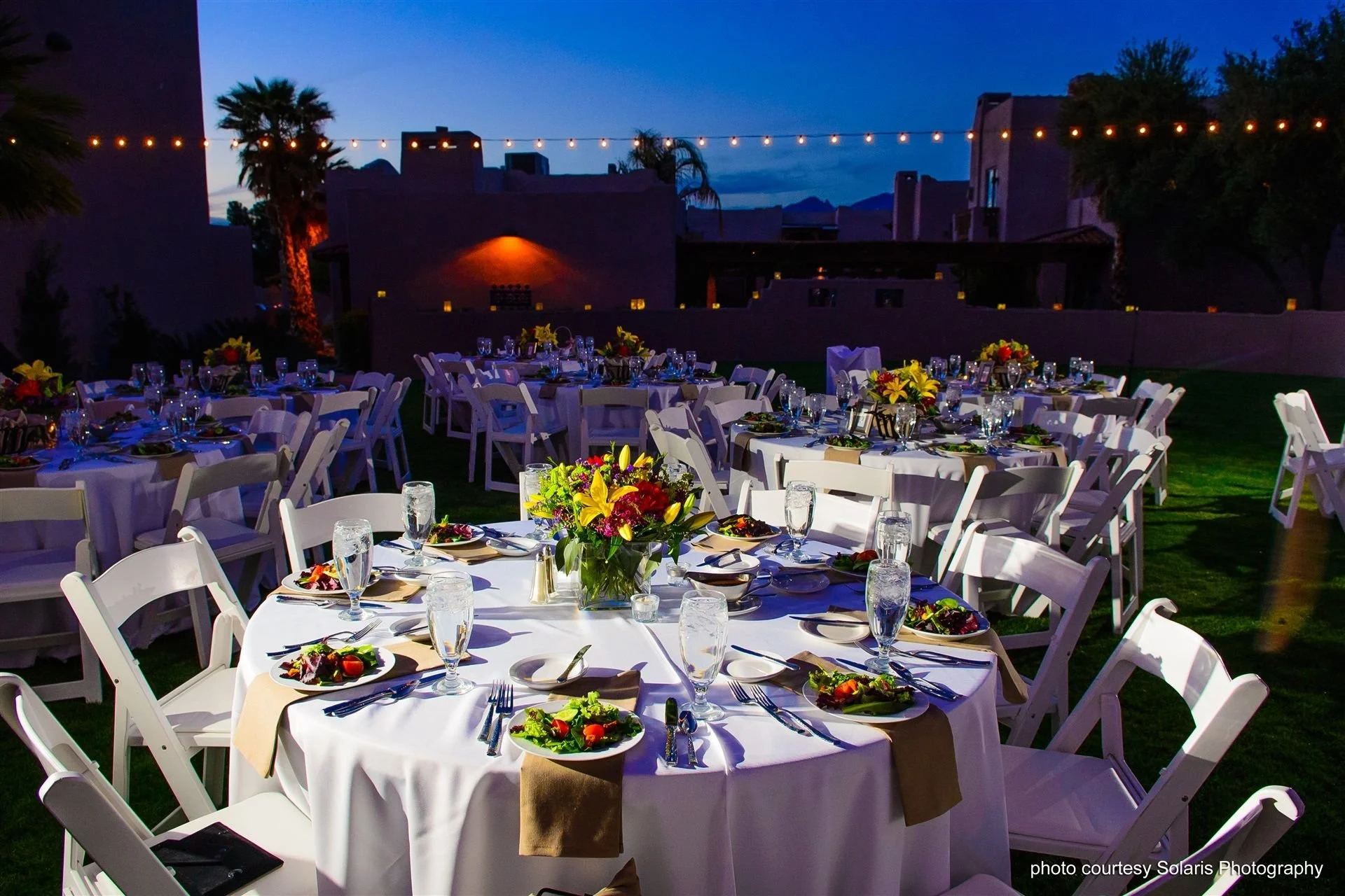 Set up for an outdoor evening event with round tables covered in white tablecloths, decorated with colorful floral centerpieces, and set with plates, glasses, and silverware. String lights are hung above, and the background features palm trees and buildings with a darkening sky.