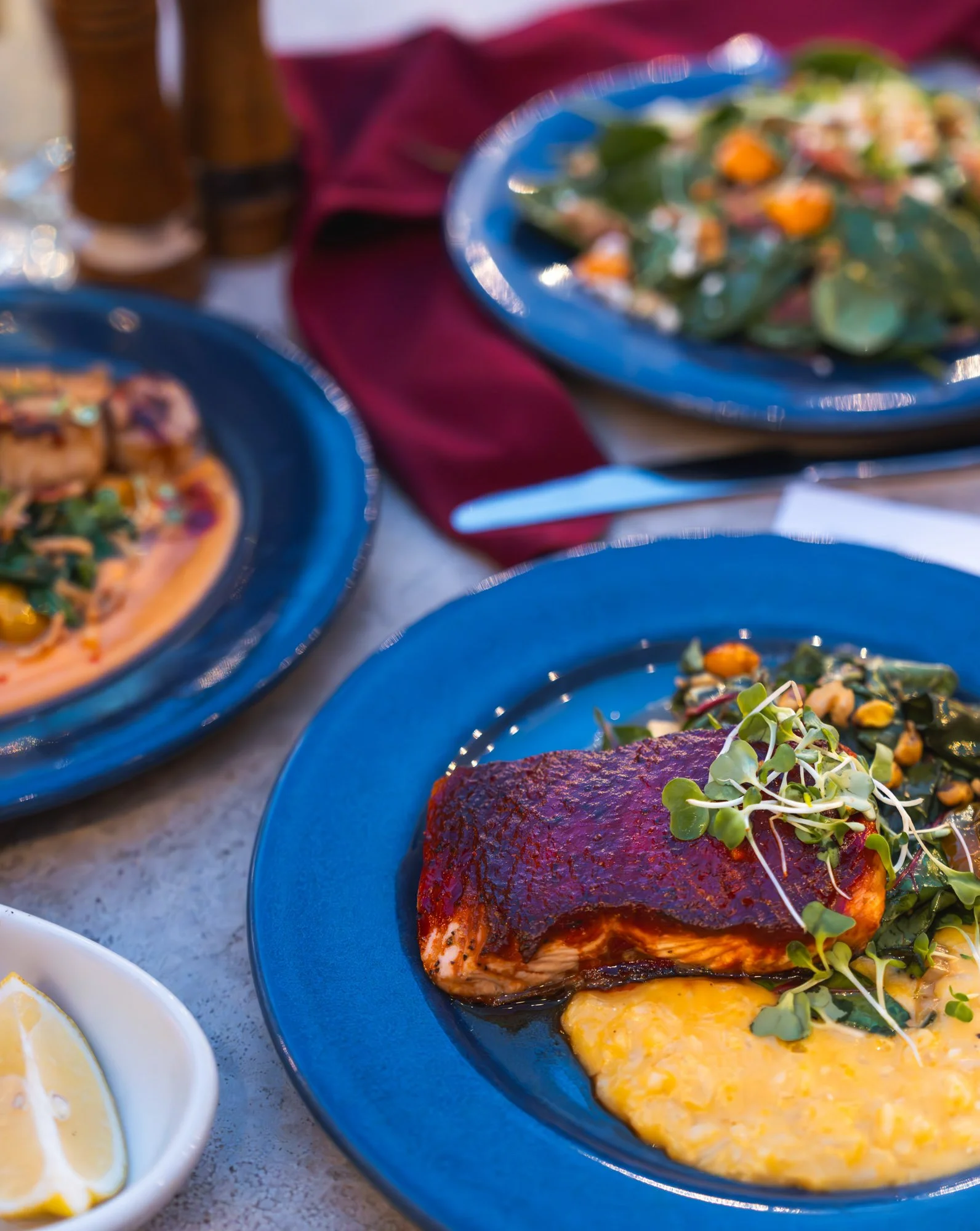Plate with salmon topped with tomato-based sauce, microgreens, and a side of scrambled eggs, surrounded by blue plates with salads on a table with lemon wedges and utensils.