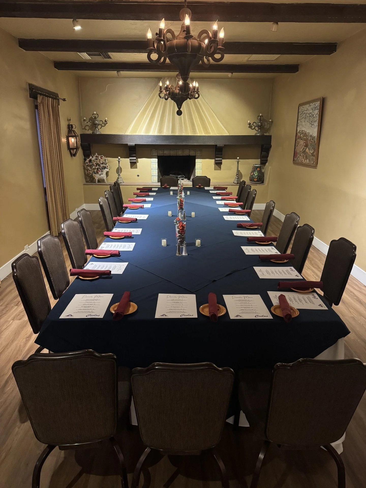 A long dining table set for a formal dinner with menus, red napkins, and gold plates in a warmly lit room with a chandelier, artwork, and a fireplace.
