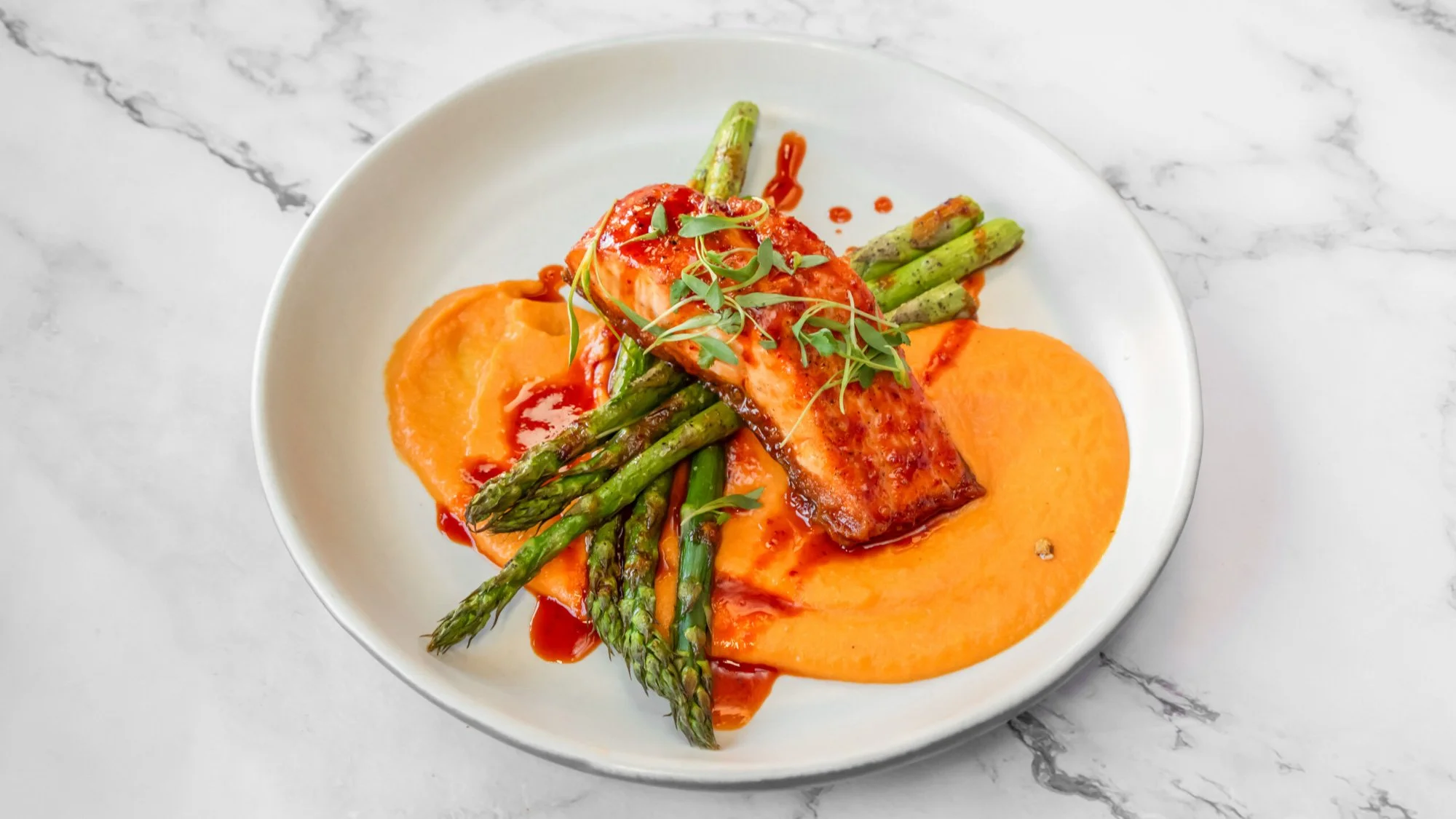 Grilled salmon topped with herbs, served with roasted asparagus, sweet potato puree, and a dollop of purée on a white plate.