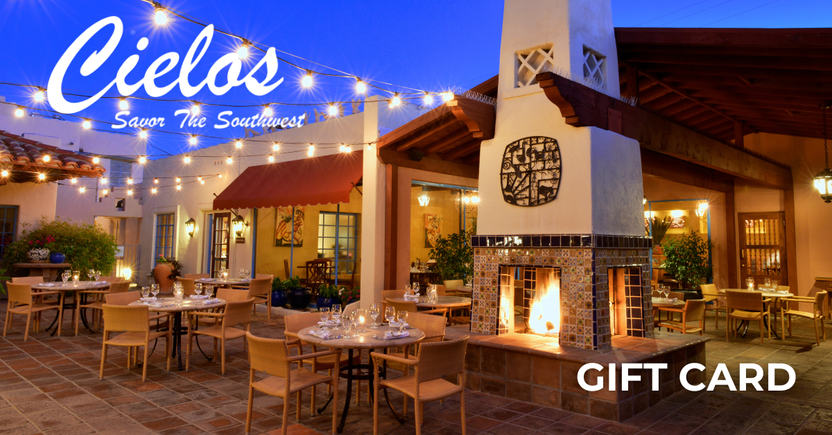 An outdoor dining area at night with tables set with glasses and silverware, string lights overhead, a large fireplace with a fire, and a sign that reads 'Cielos Savor The Southwest'.