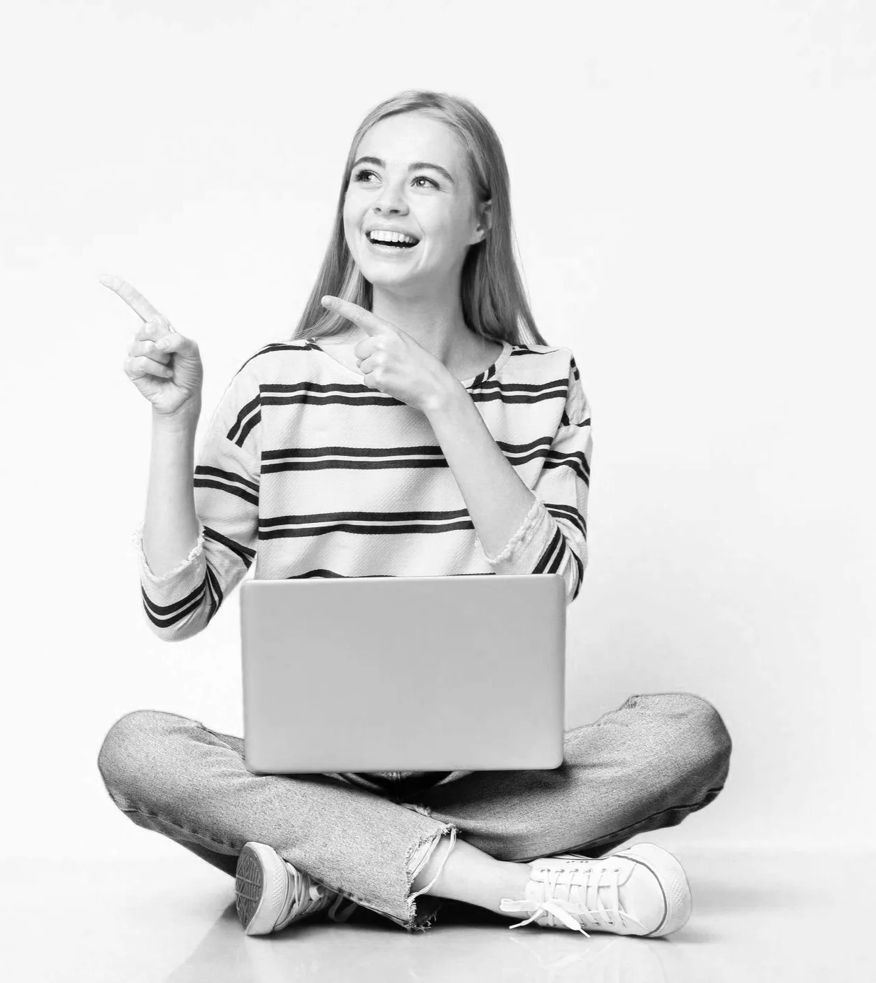 A woman sitting cross-legged on the floor with a laptop on her lap, smiling and pointing to her left.
