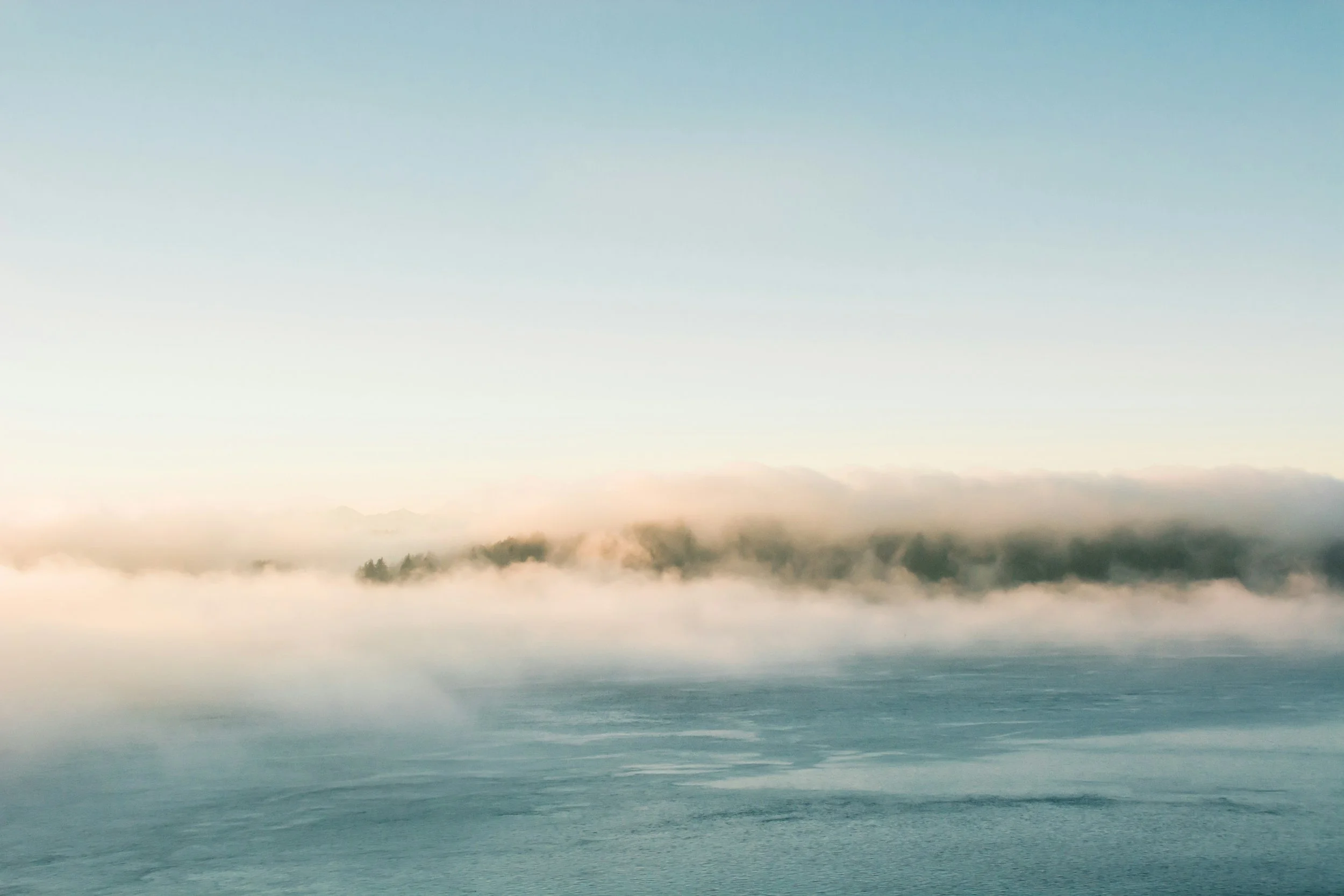 Calm ocean water with a foggy shoreline and distant hills under a clear sky.