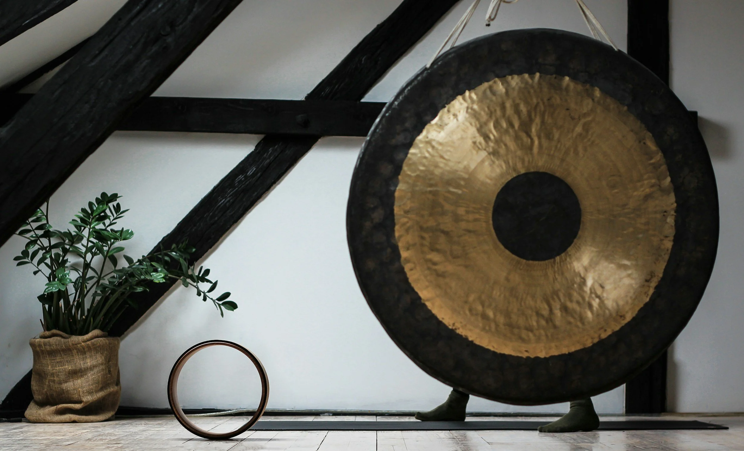 Large gong with a golden center and black outer rim, standing on feet, placed in a room with wooden beams and a potted plant in a burlap sack.