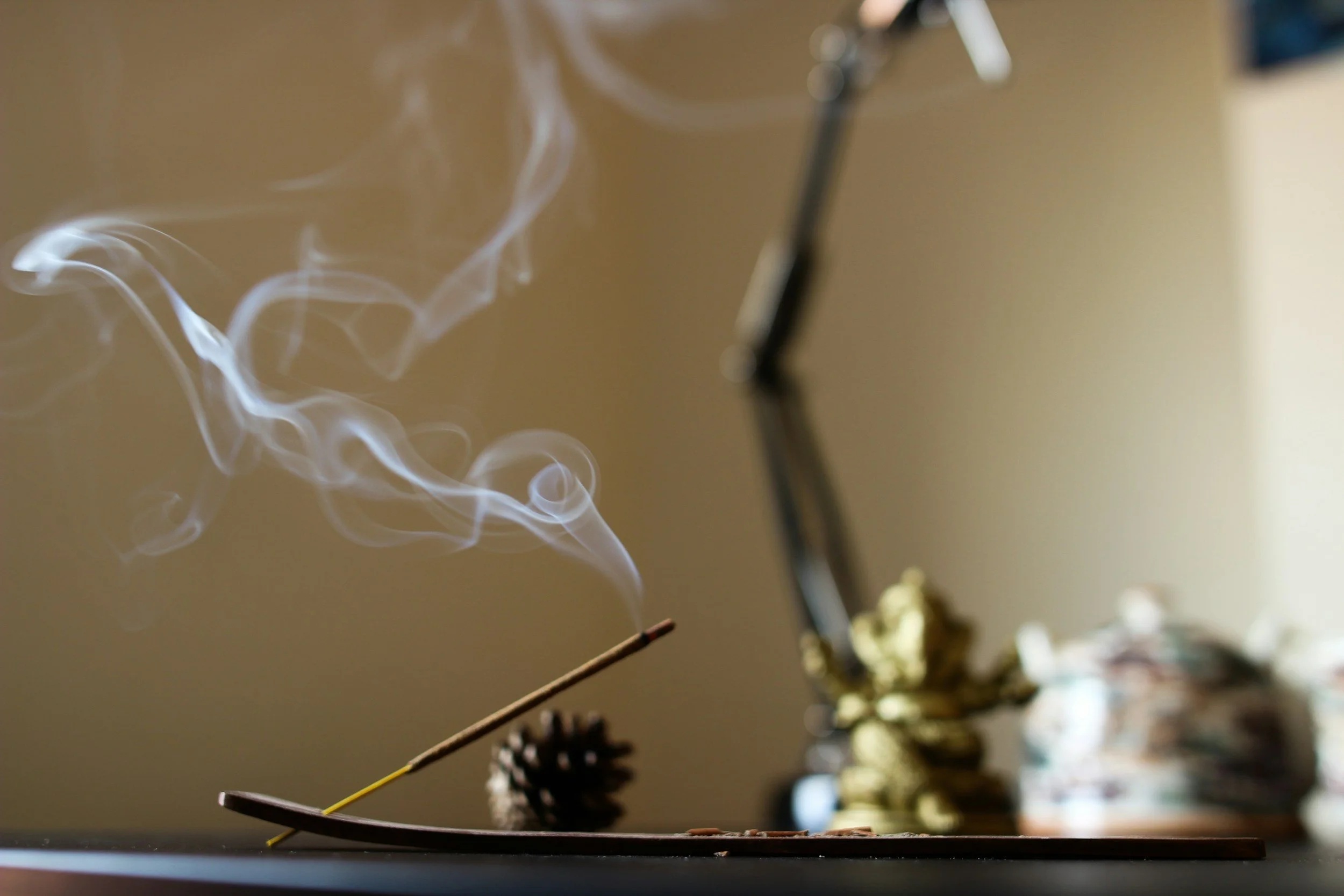 A lit incense stick on an incense holder emitting swirling smoke, with a blurred background including pinecones and a decorative teapot.