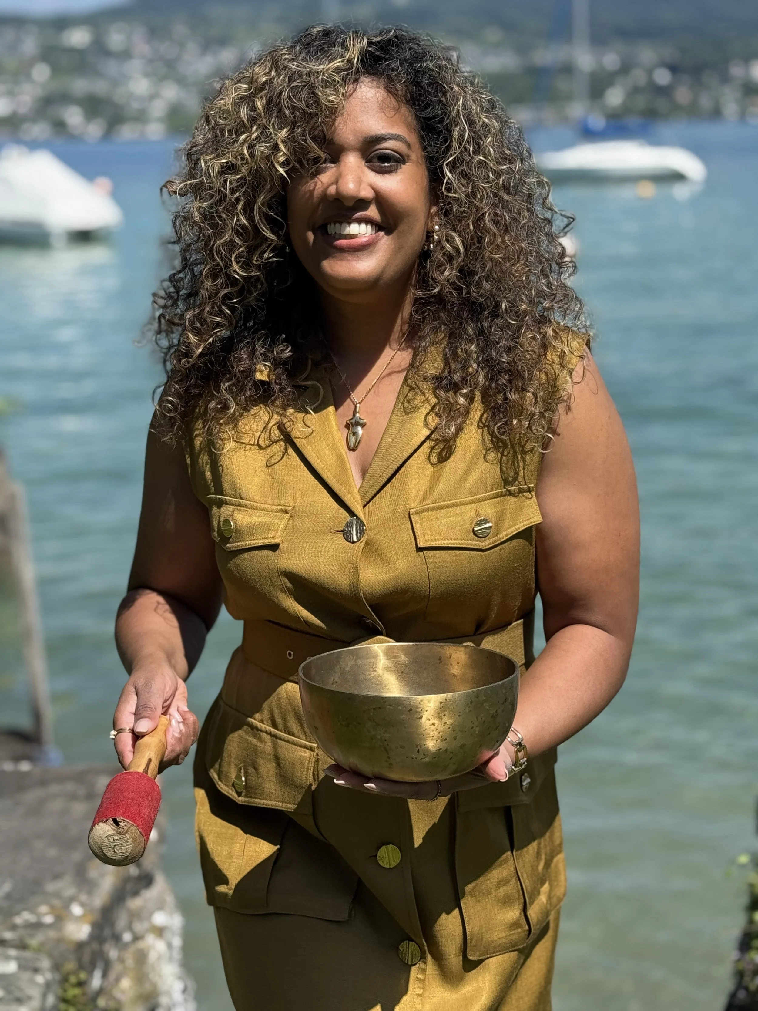 Woman with curly hair smiling, holding a singing bowl in her left hand and a mallet in her right hand, outdoors near a body of water with boats in the background.