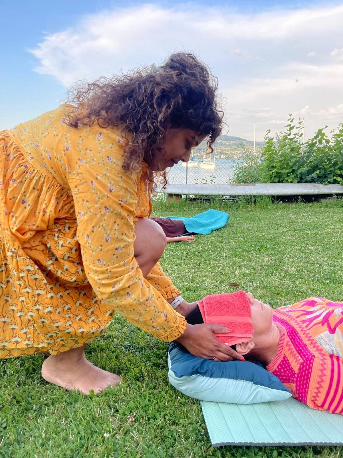 A woman practicing cervical spine yoga massage on a woman lying on a mat outdoors near a body of water with sailboats in the background.