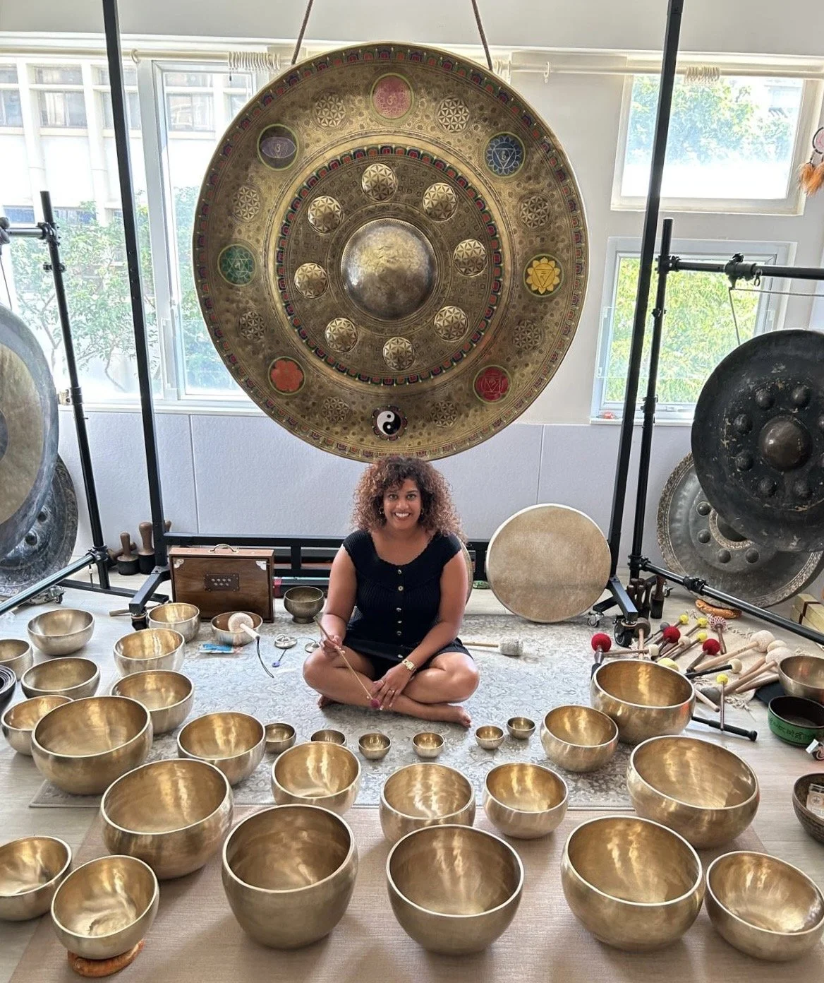 A woman sitting cross-legged on the floor surrounded by brass singing bowls of various sizes. Behind her are large gongs and a large decorative cymbal hanging above. The scene appears to be in a well-lit room with windows showing greenery outside.