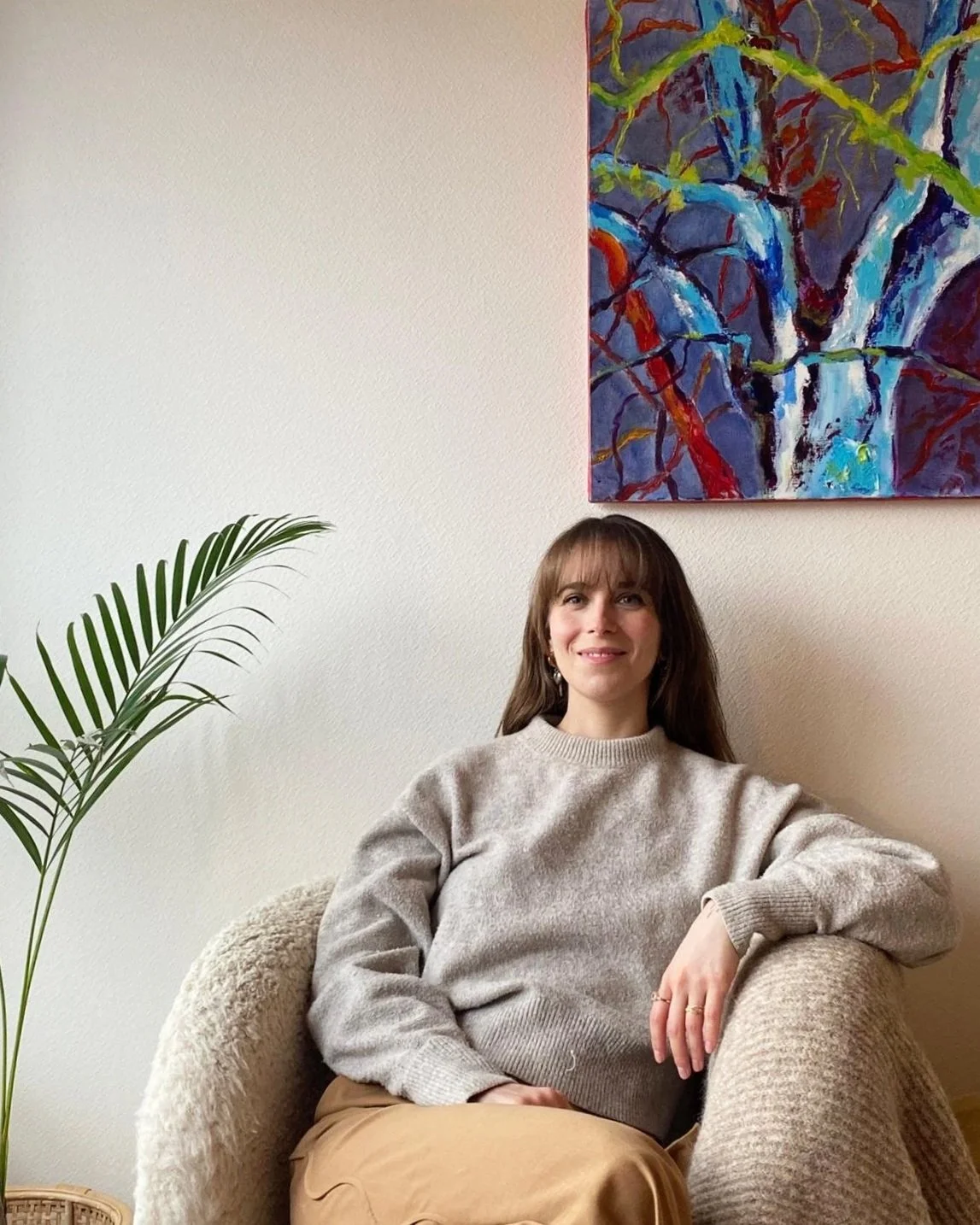 Une femme assise dans un fauteuil en peluche beige, portant un pull gris clair et une jupe beige, souriant vers l'appareil, avec une peinture abstraite colorée suspendue au mur derrière elle et une plante verte à gauche.