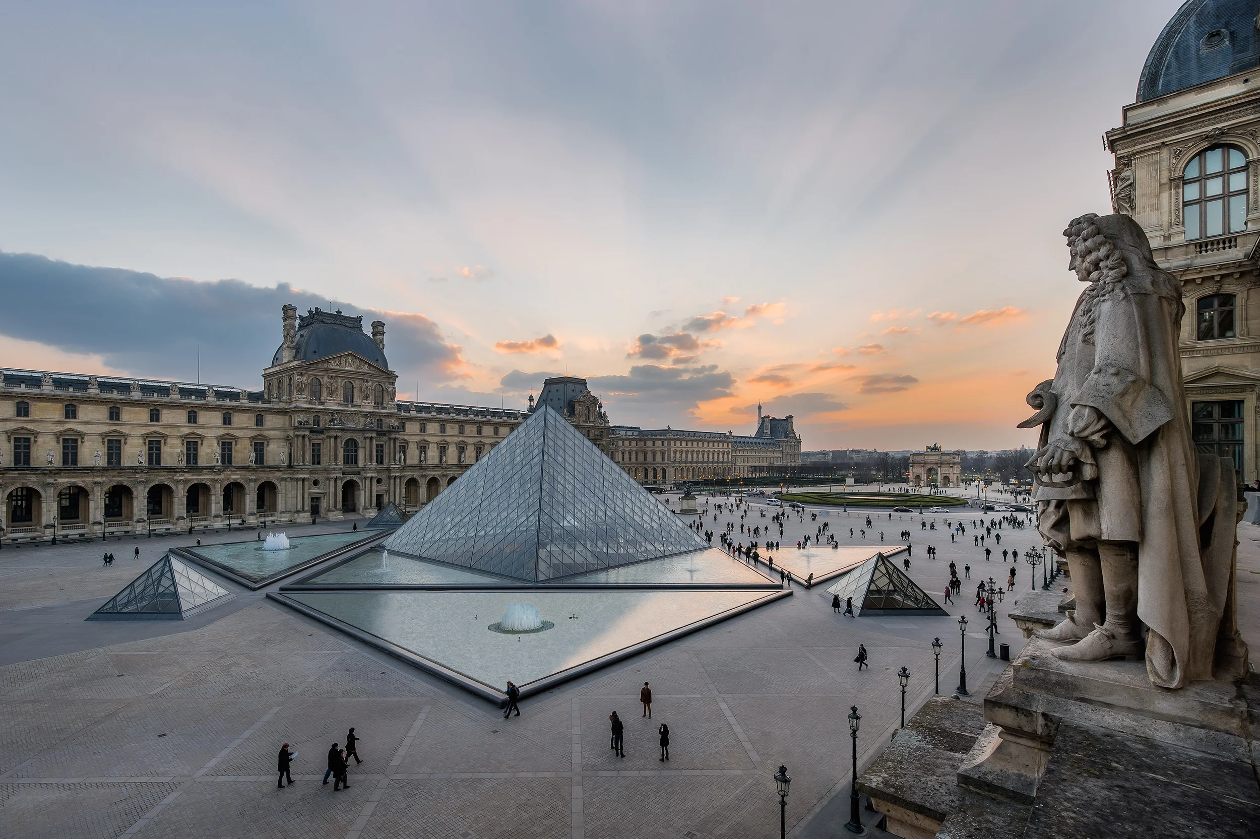 Vue de la cour du Louvre avec les pyramides en verre au coucher du soleil, statues en pierre, et un ciel nuageux.