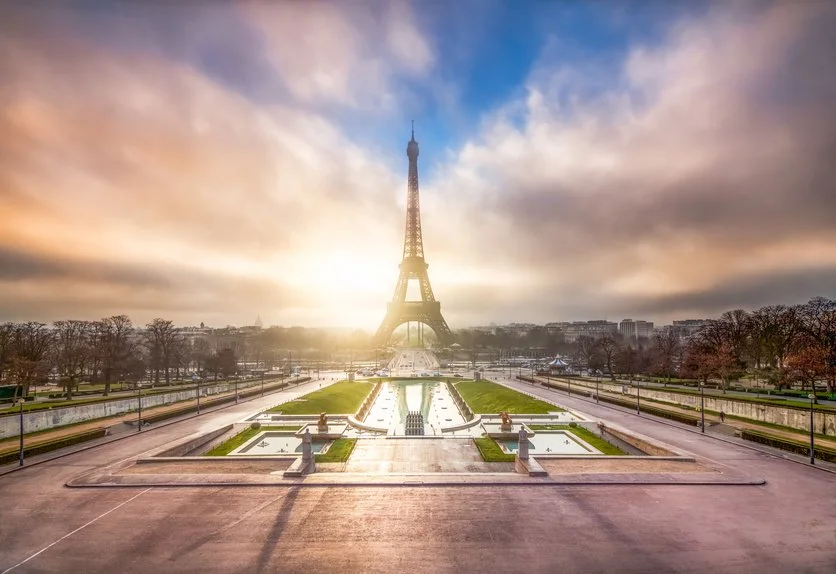 La Tour Eiffel au lever ou coucher du soleil avec le ciel nuageux, vue depuis le Champ de Mars avec des fontaines et jardins.